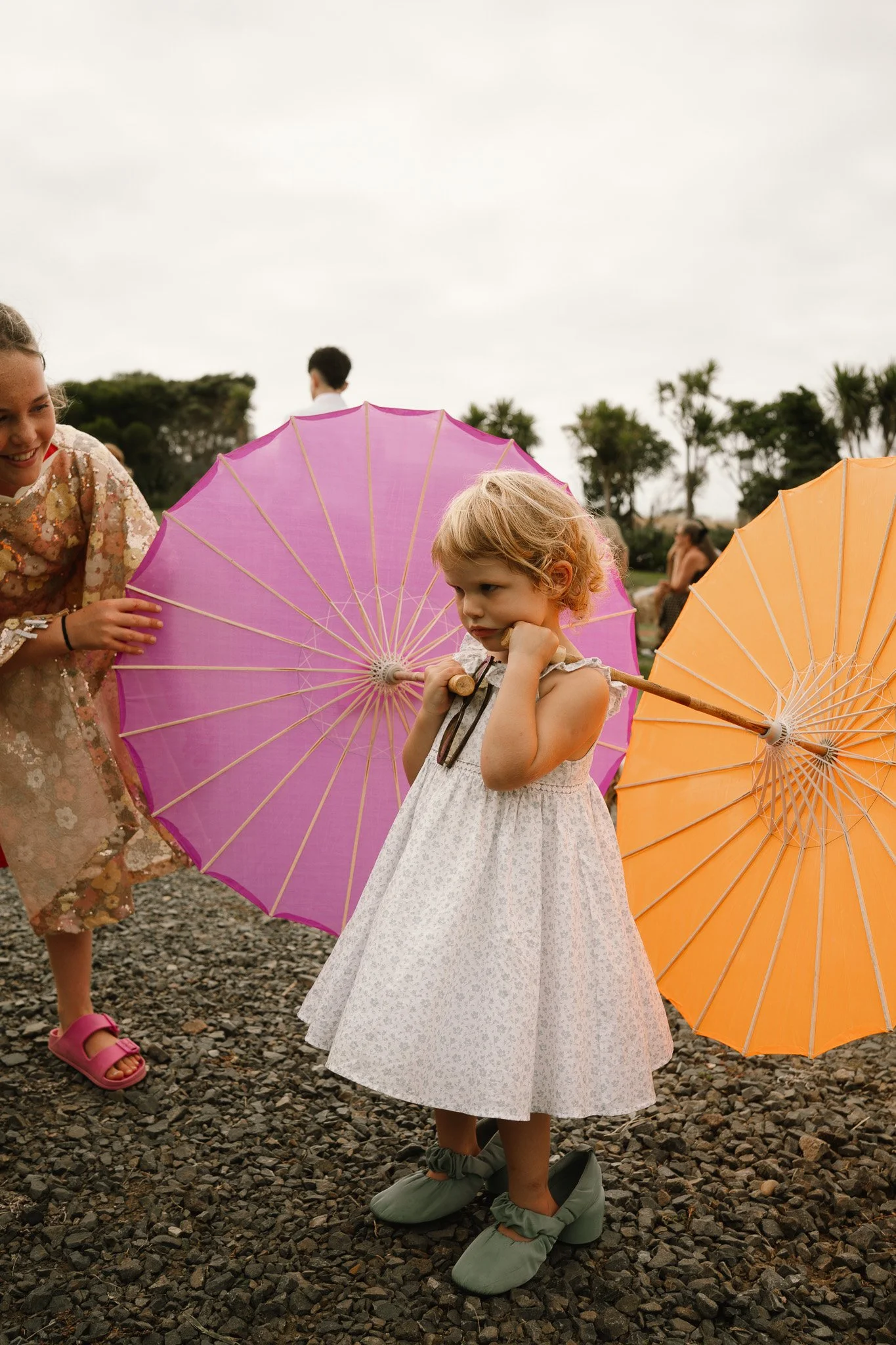 A young girl with blonde hair in a white dress holding an orange umbrella. An older girl in a floral dress and pink sandals appears to be adjusting a pink umbrella behind her. There are trees and other people in the background on a cloudy day.