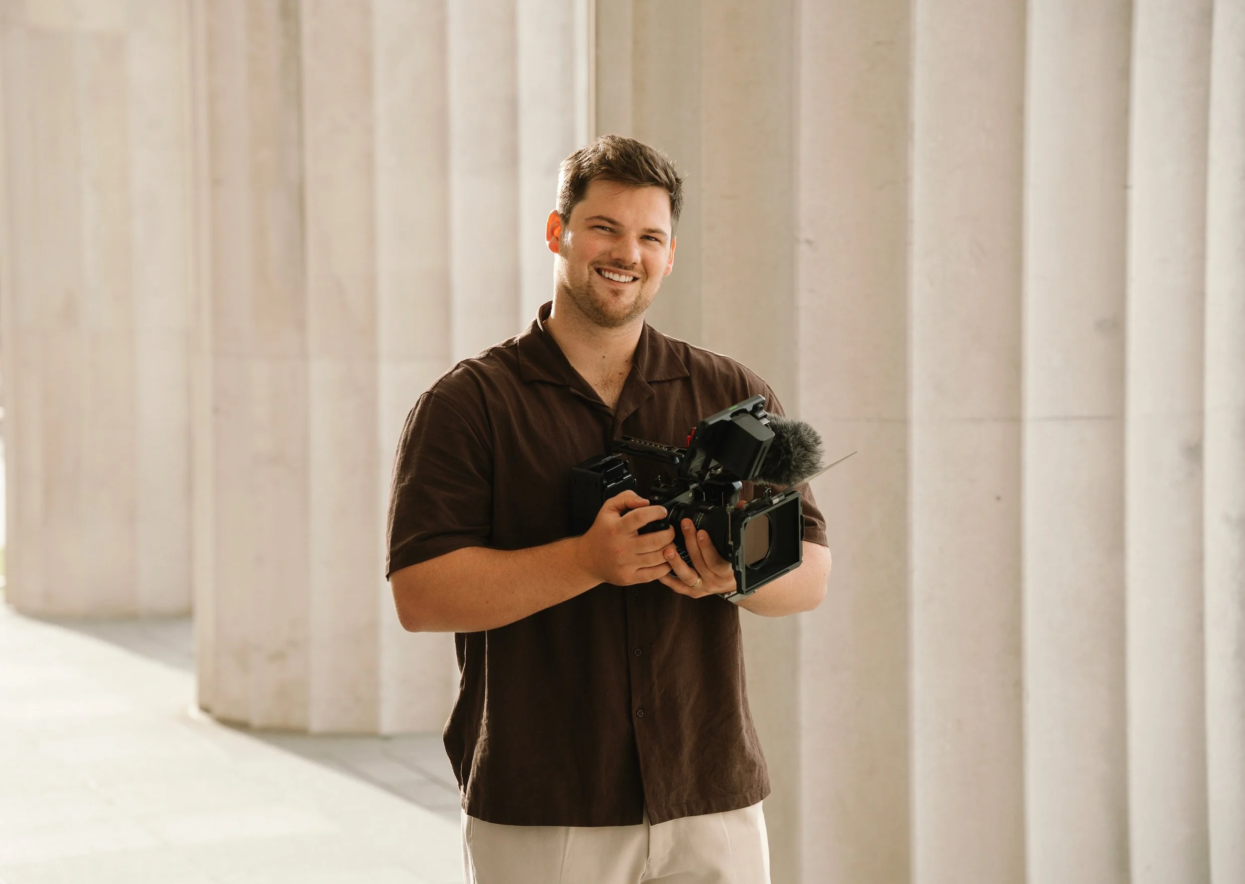 A smiling man holding professional video camera equipment stands outdoors against a background of large concrete columns.