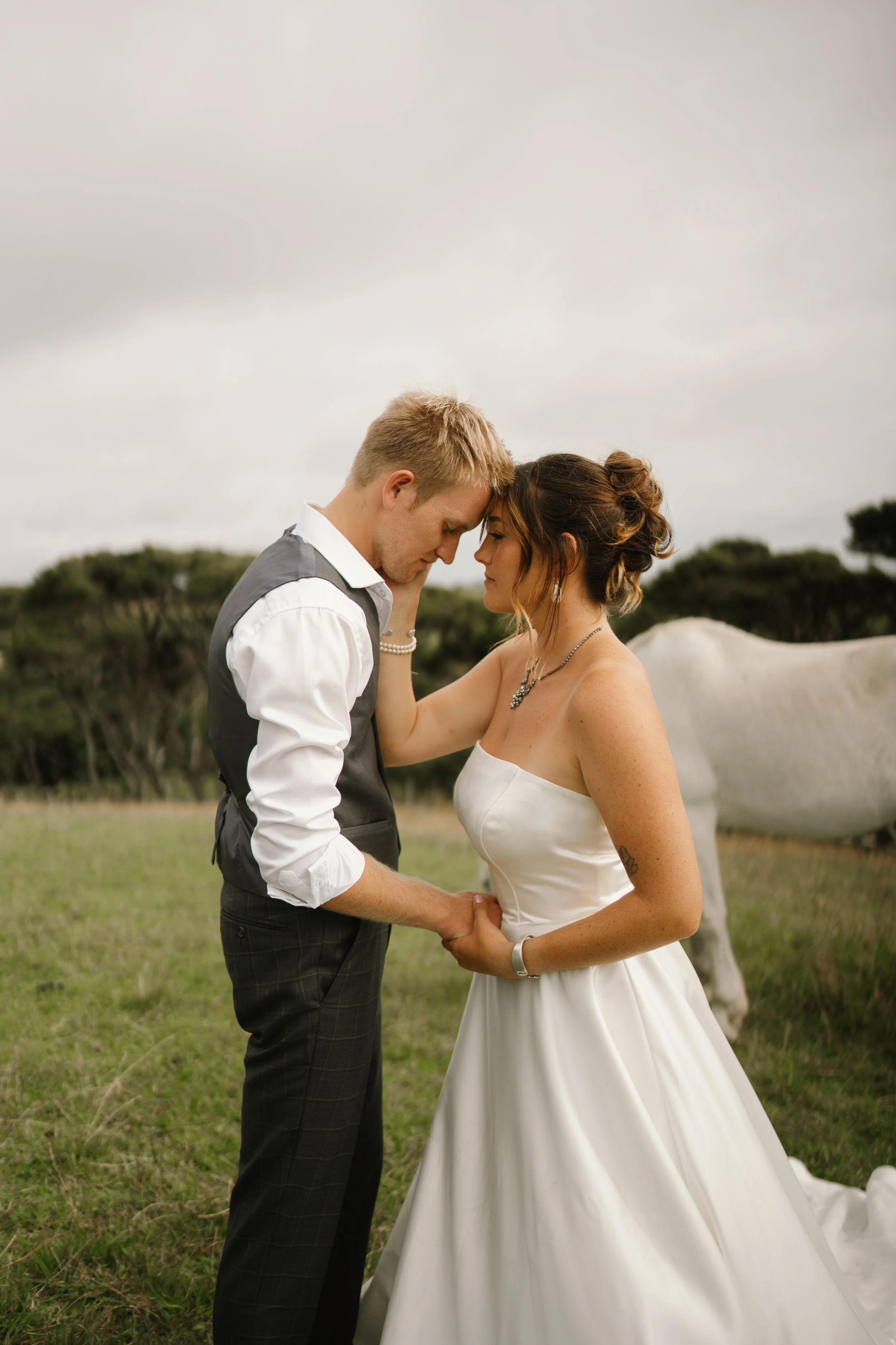 A newlywed couple standing close together outdoors, foreheads touching, holding hands, with a white horse in the background.