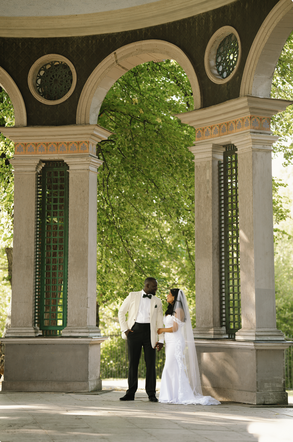 Bride and groom holding hands under a decorative pavilion with green trees in the background.
