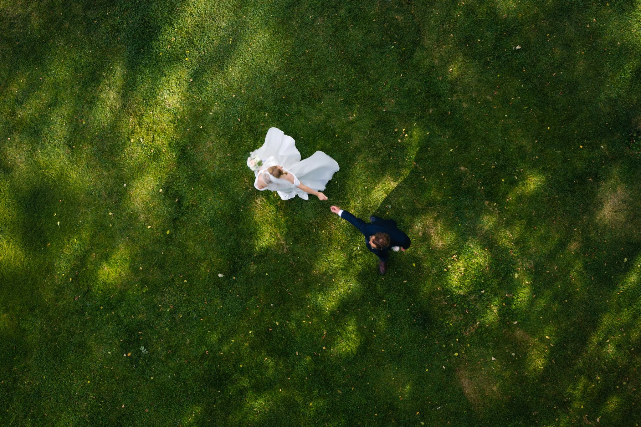 A bride in a white wedding gown holding hands with a groom in a dark suit, walking on green grass in an outdoor setting.