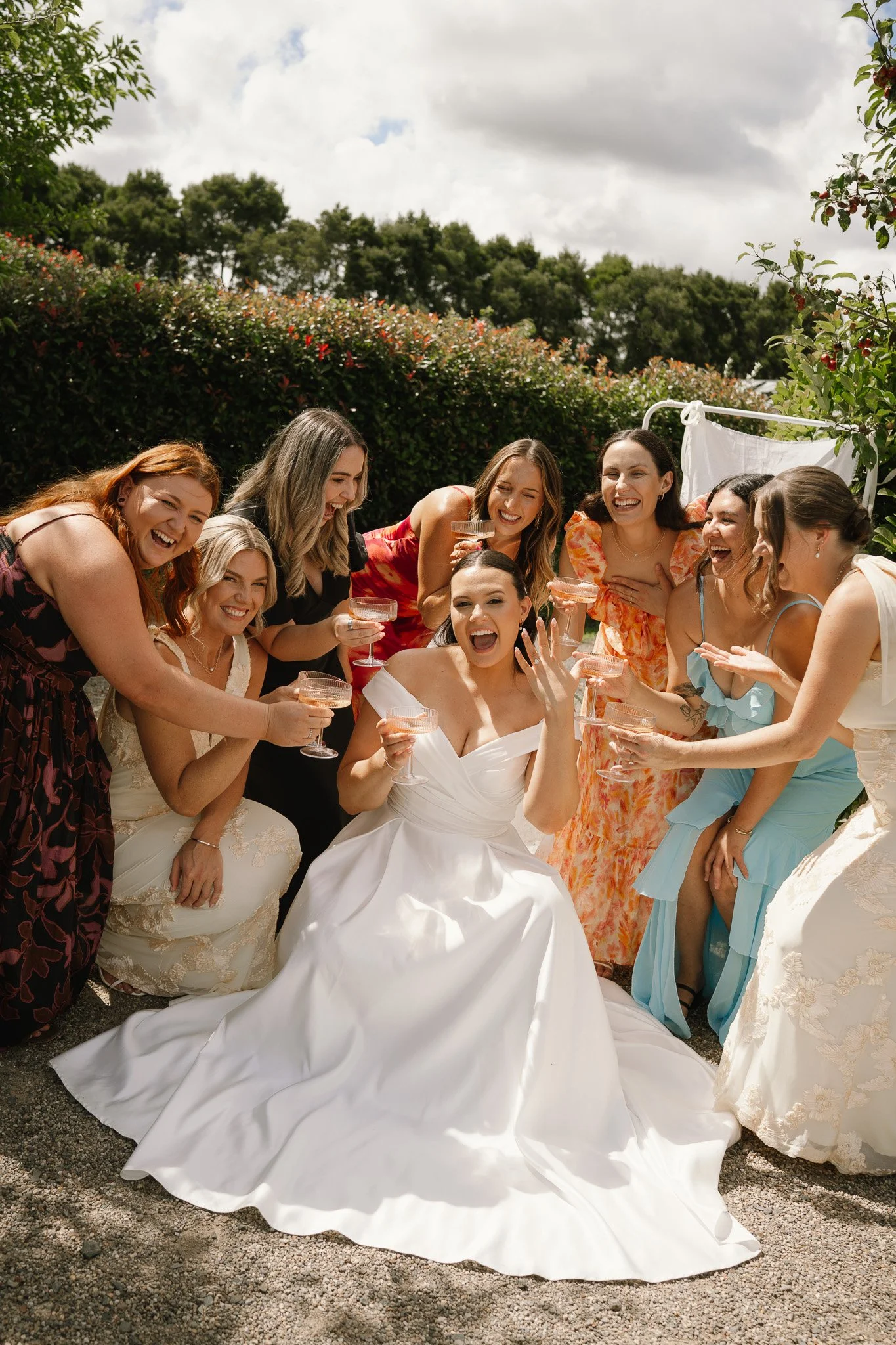 A bride and her friends celebrating outdoors, dressed in elegant gowns, holding glasses of champagne, and smiling joyfully.