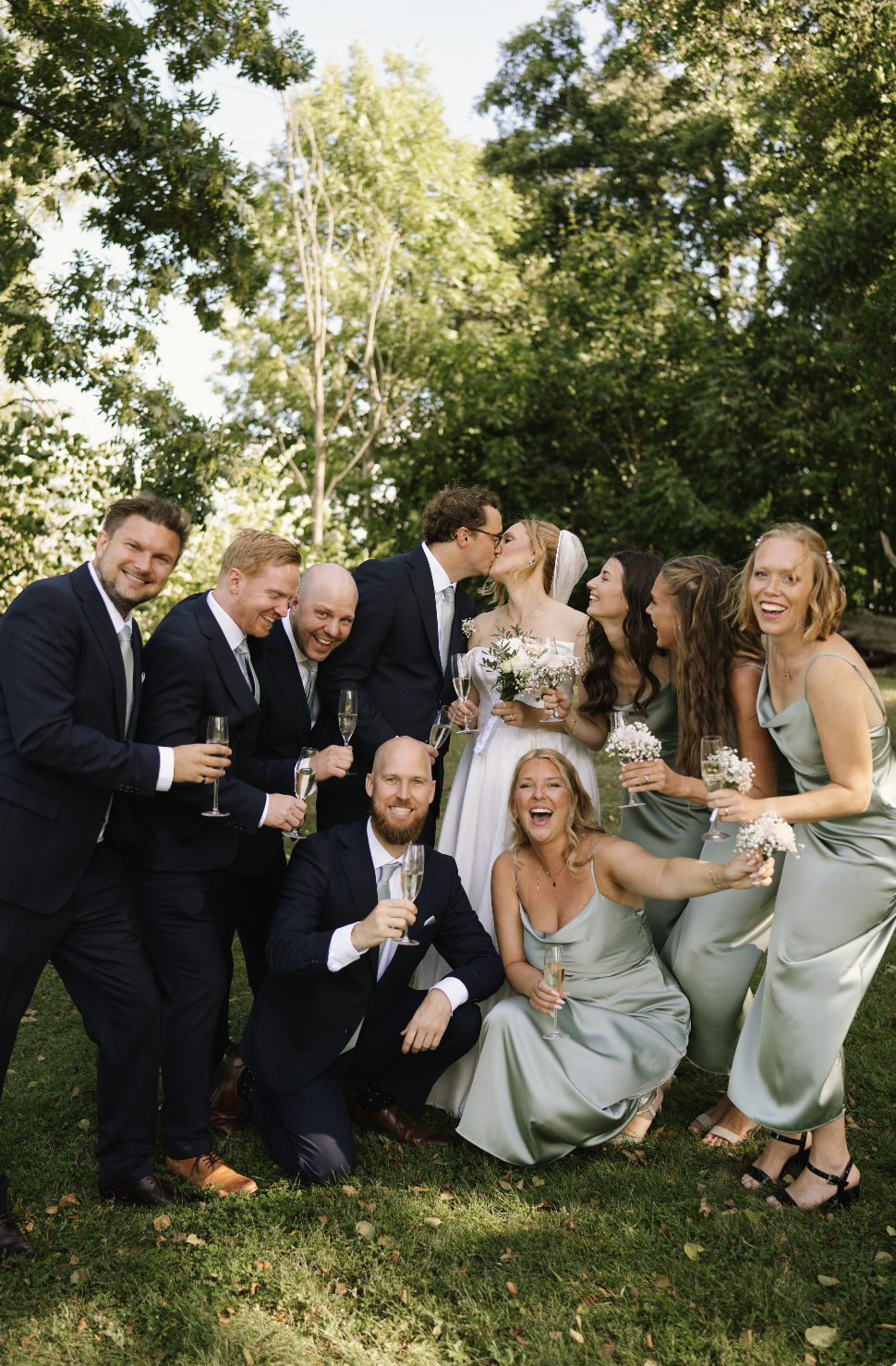 A group of wedding party members, including bride and groom, celebrating outdoors with glasses of champagne, in a green, wooded setting.