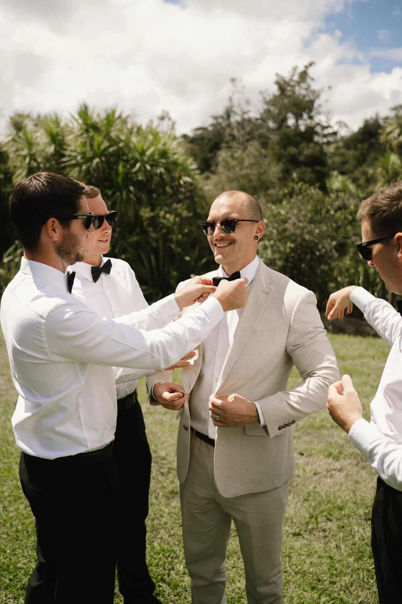 Group of five men in formal attire, some adjusting bow ties on a groom, outdoors with green trees and cloudy sky in background.