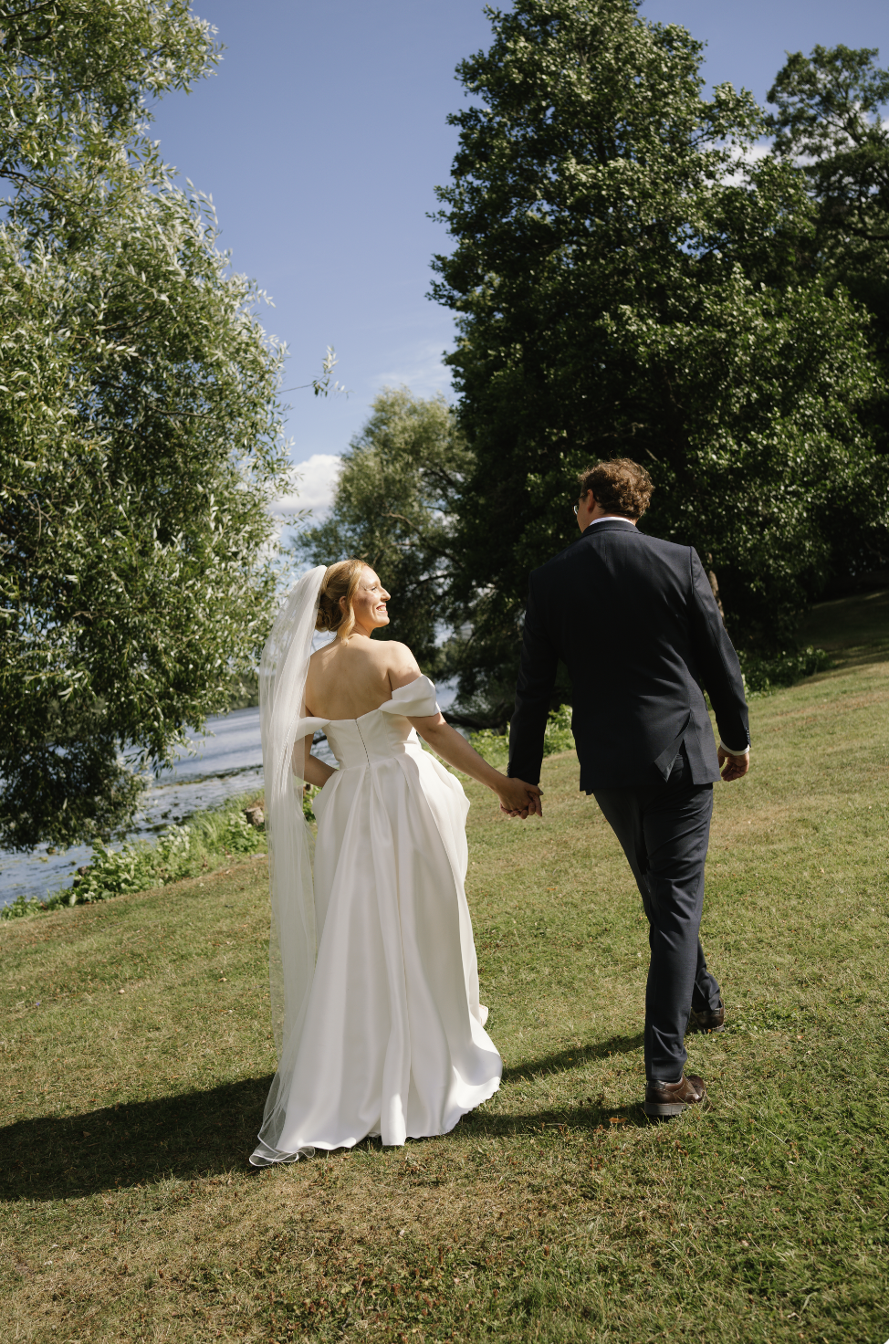 A bride and groom walking hand-in-hand outdoors near a river, surrounded by trees and a clear blue sky.