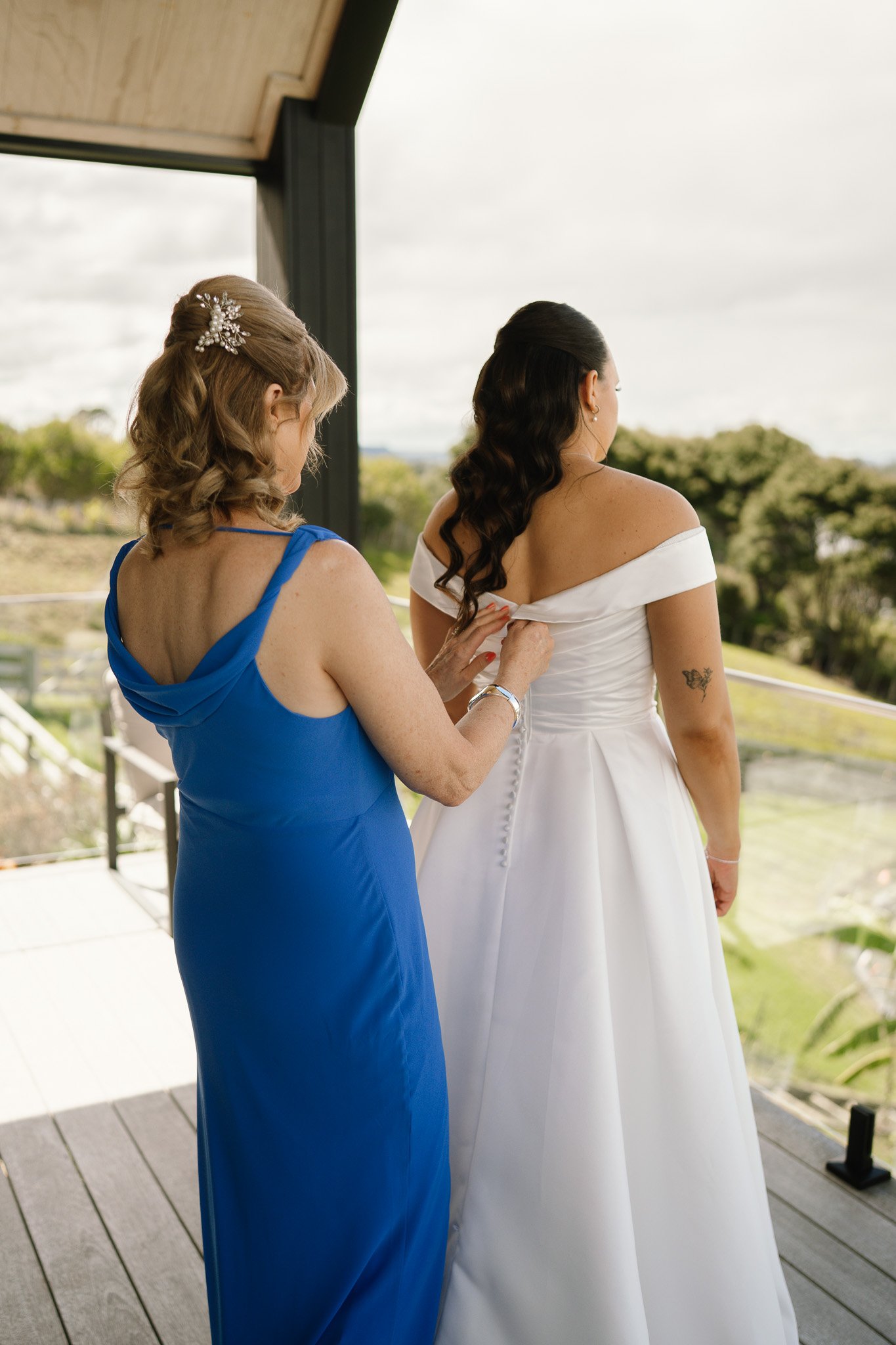 A woman in a white wedding dress is being helped with the back of her dress by another woman in a blue dress, on a balcony overlooking a garden.