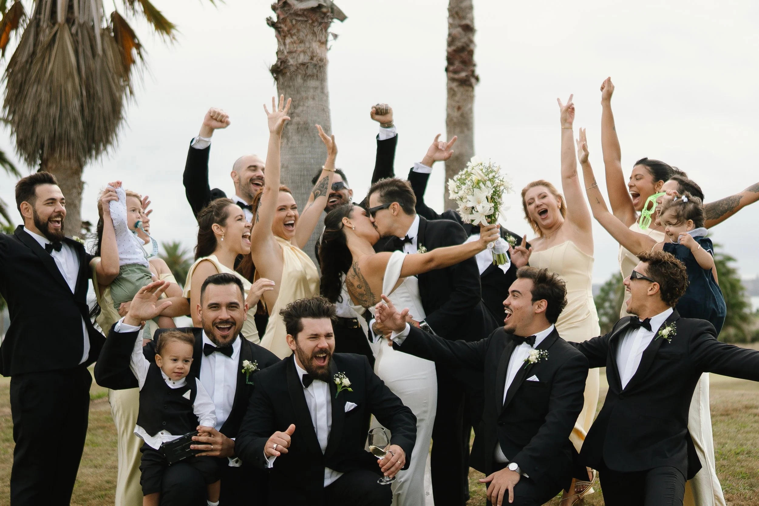 A group of people in formal attire celebrating outdoors, with two women in the center kissing, one holding a bouquet, surrounded by smiling individuals, palm trees, and a cloudy sky.
