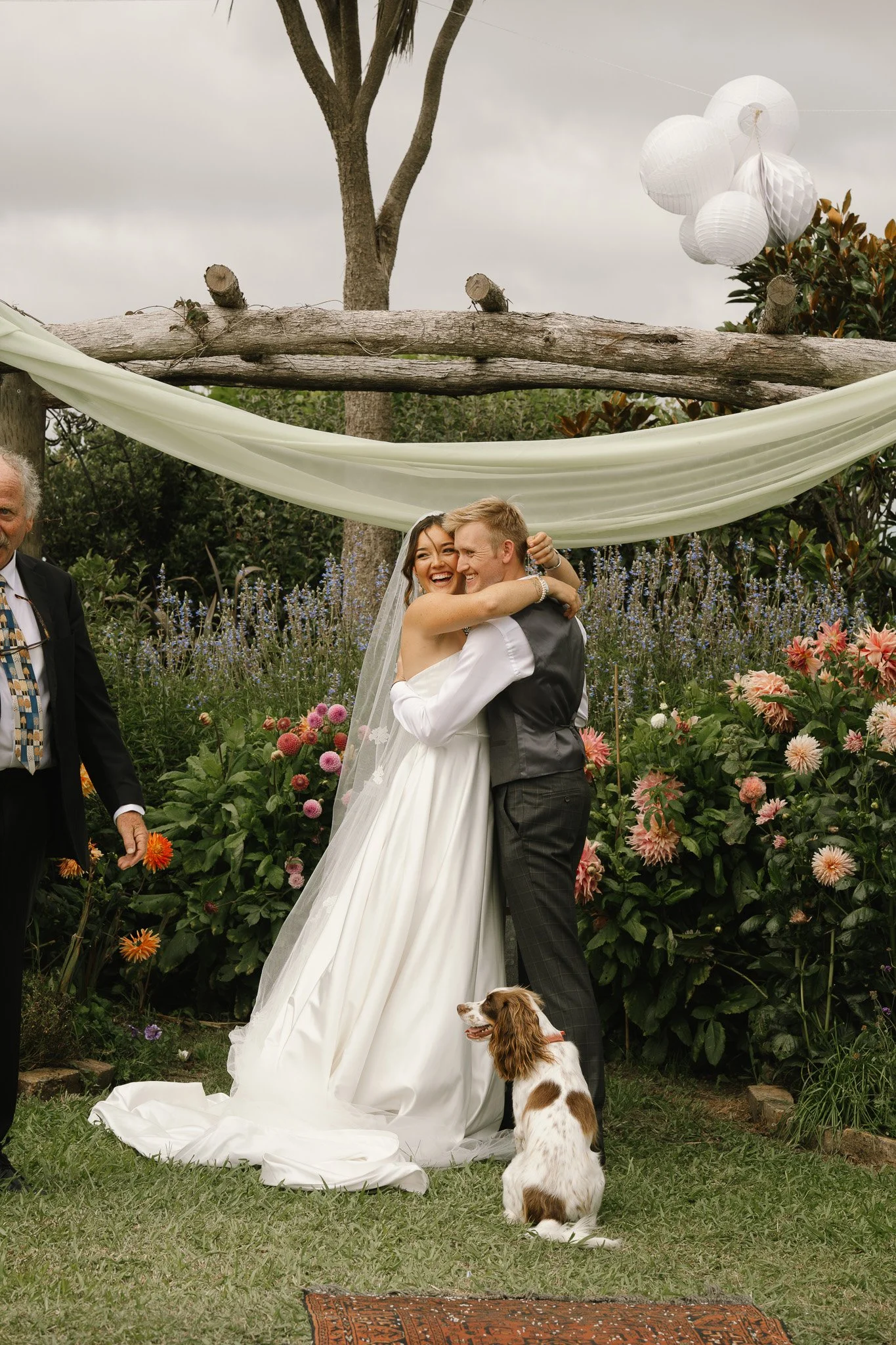 A bride and groom embrace during their wedding ceremony in an outdoor garden, surrounded by flowers, with a dog sitting on the grass nearby, and decorative lanterns hanging overhead.