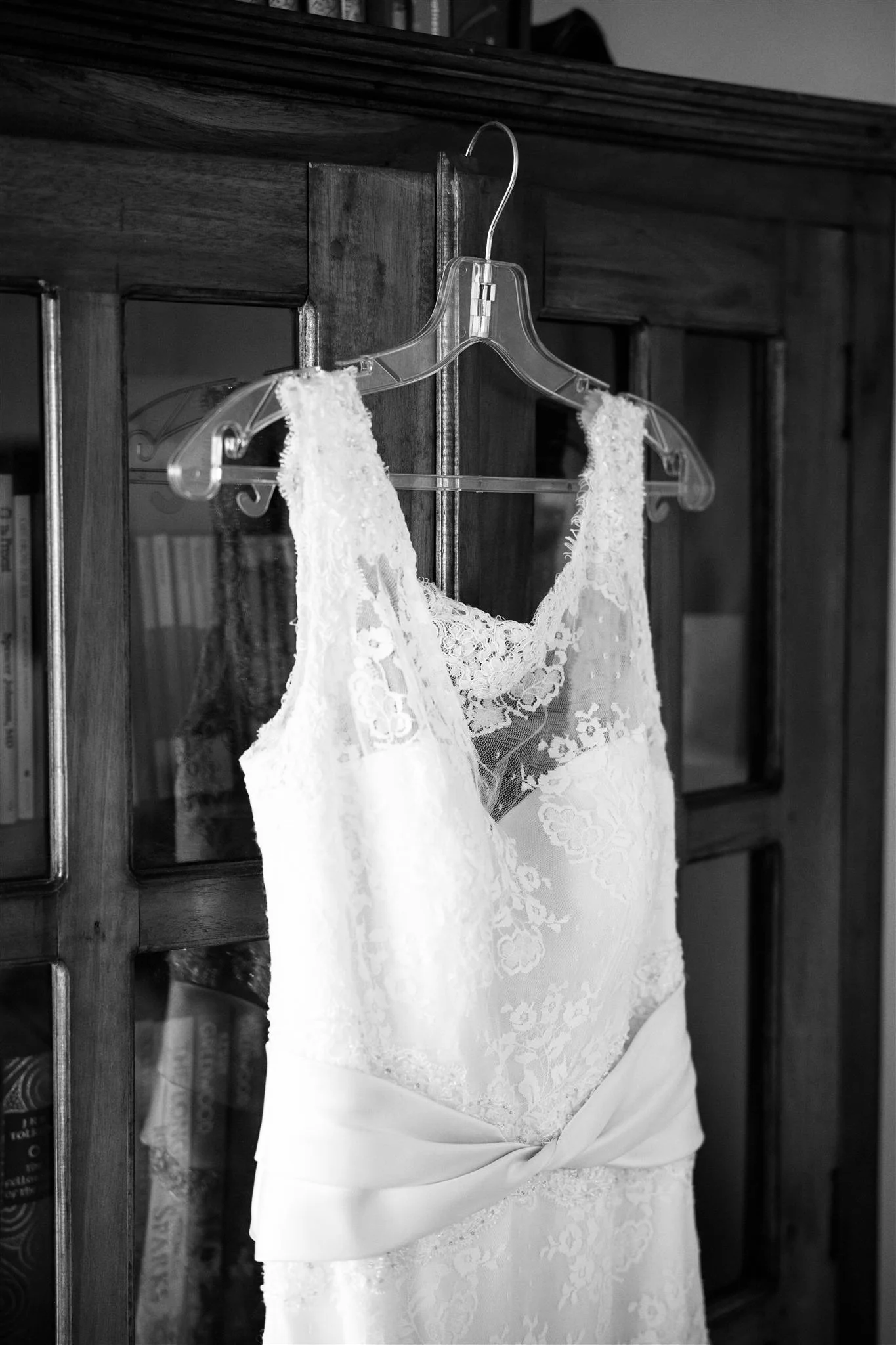 A wedding dress with lace details hanging on a clear plastic hanger on a wooden cabinet door.