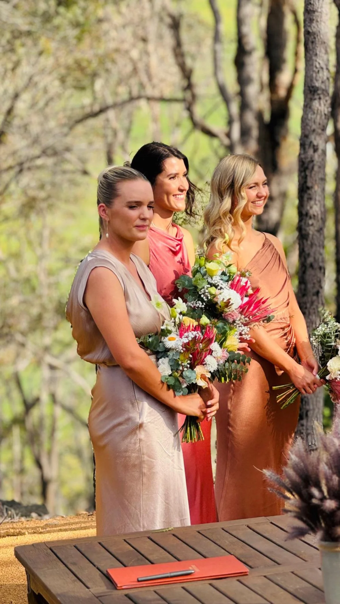 Three bridesmaids in elegant dresses standing outdoors in a wooded area, holding bouquets of flowers, participating in a wedding.
