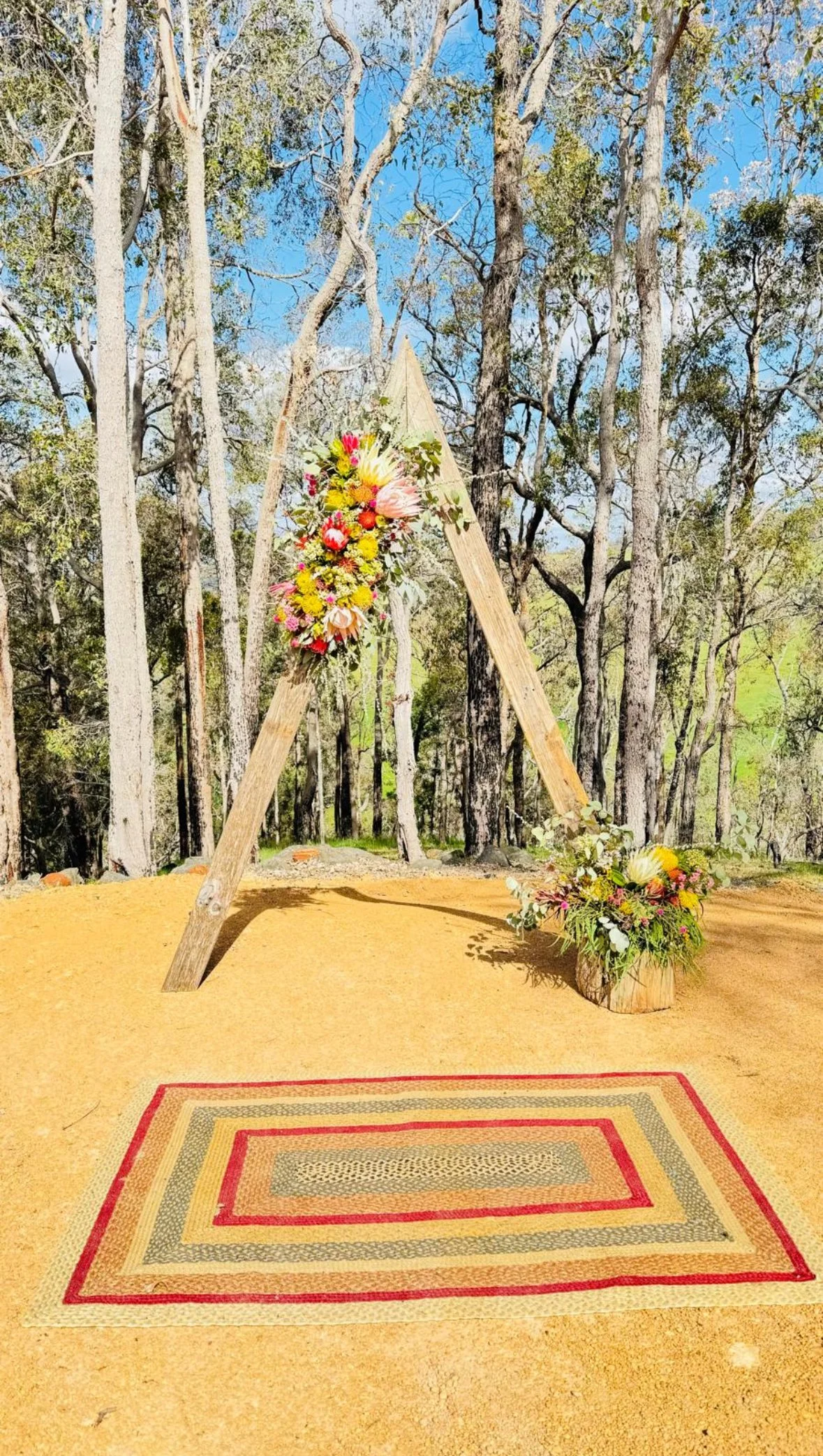 A wooden arch decorated with colorful flowers stands outdoors on a dirt area, with a patterned rug on the ground and trees in the background.
