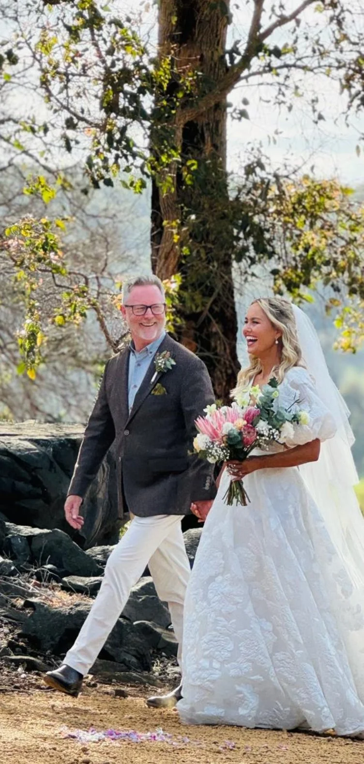 A bride and her father walking outdoors during a wedding ceremony, holding hands and smiling, with trees and a natural setting in the background. The bride is holding a bouquet of pink and white flowers.