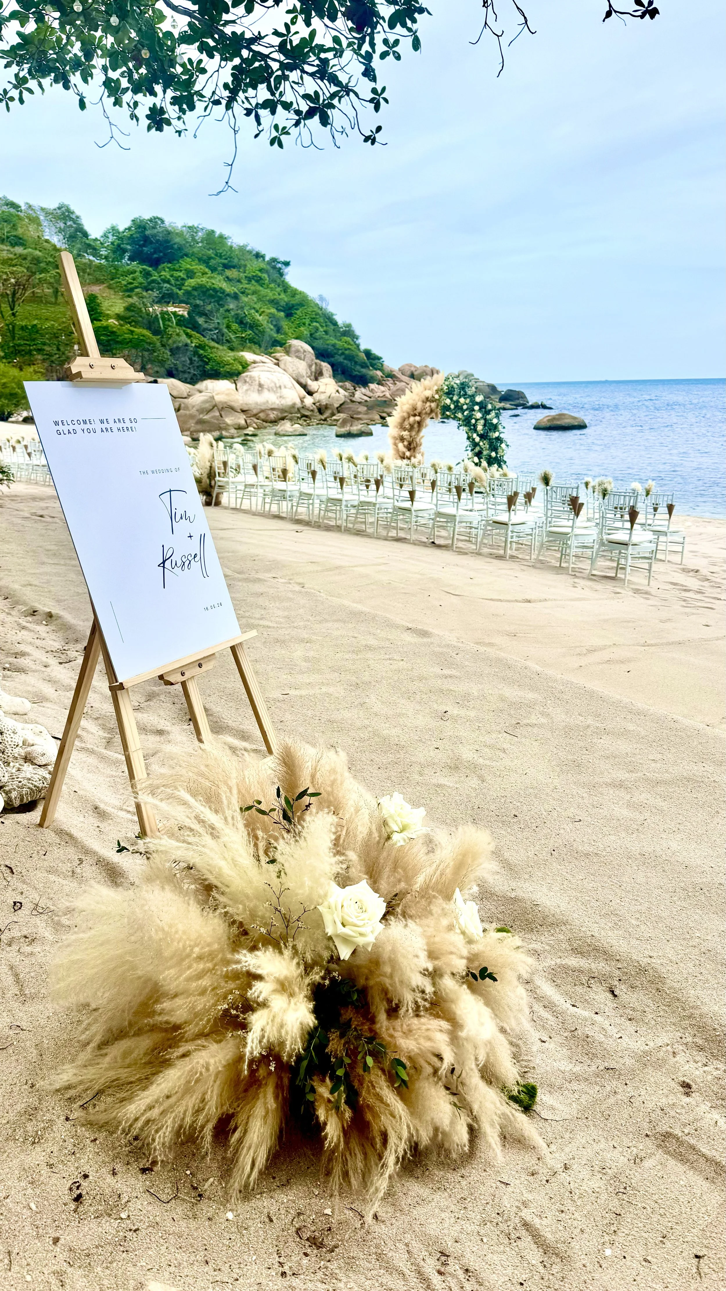 Wedding setup on a beach with white chairs and floral decorations near the shoreline, a welcome sign with wedding details, and a floral arrangement with pampas grass and white roses in the foreground.