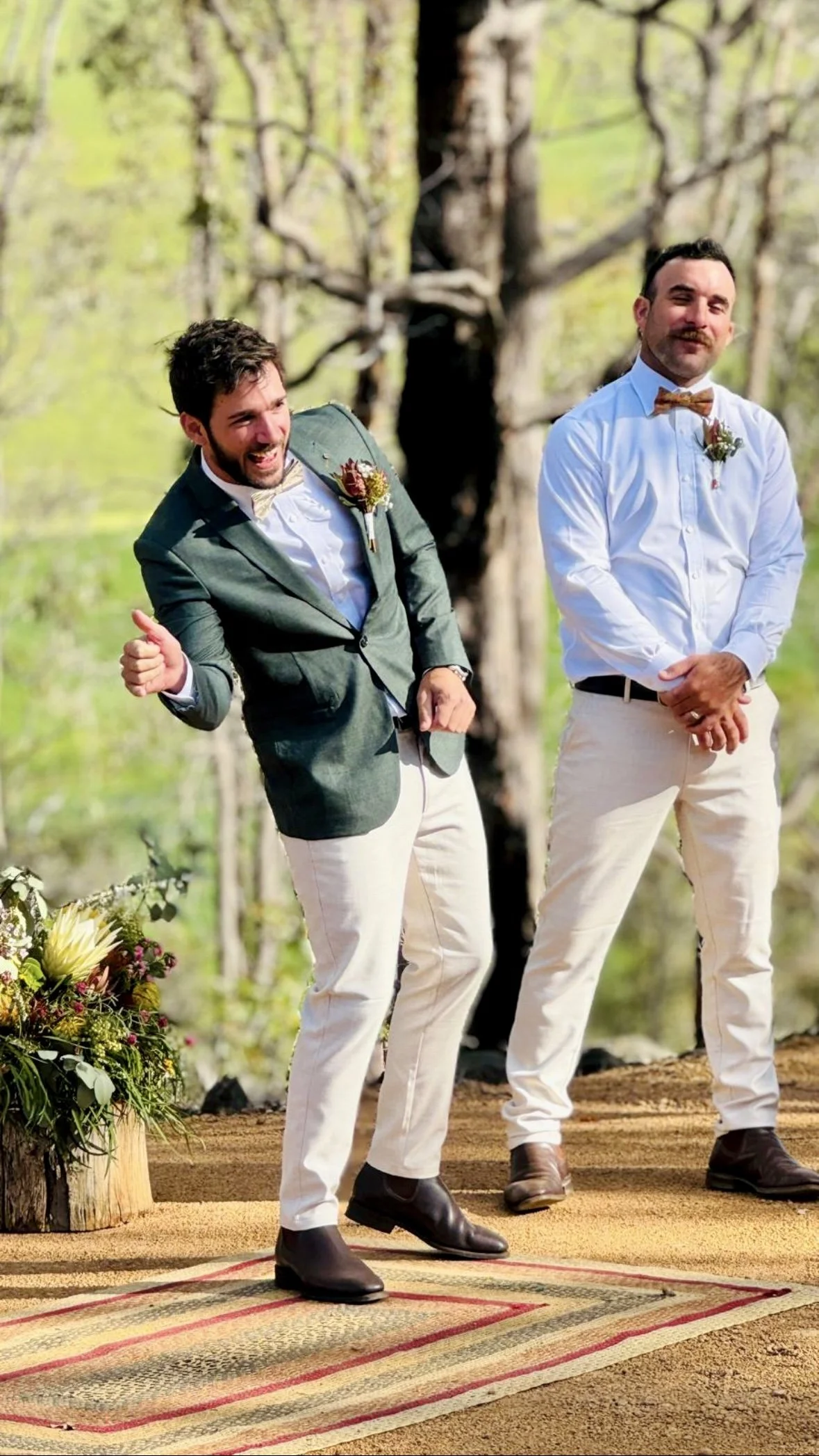Two men dressed in formal attire stand outdoors in a wooded area, likely during a wedding ceremony. One man is wearing a green jacket with a boutonnière, a white shirt, cream-colored pants, and dark boots, giving a thumbs-up and smiling. The other ma