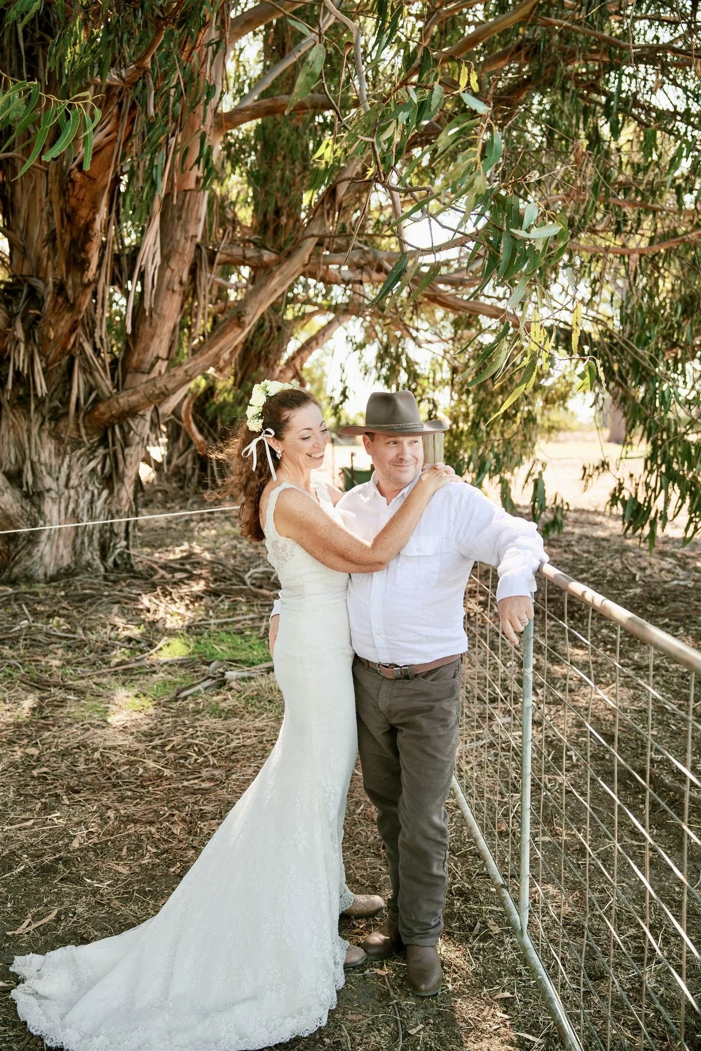 A bride in a white lace wedding dress and floral headpiece smiling, standing with her groom in a white shirt, brown pants, and a ranger hat, near a large tree and a wire fence in a natural outdoor setting.