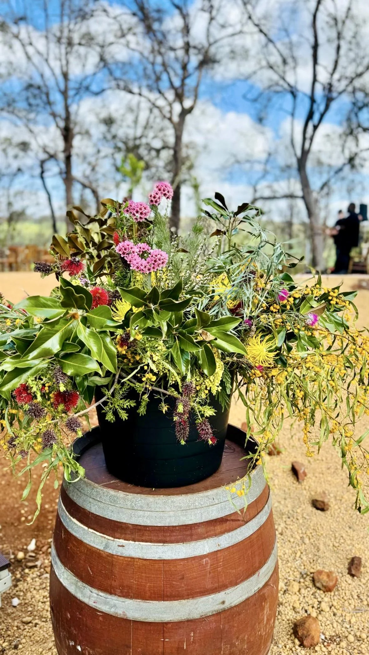  colorful flowers and green leaves placed on top of a decorated wine barrel outdoors, with a background of trees and a blue sky.