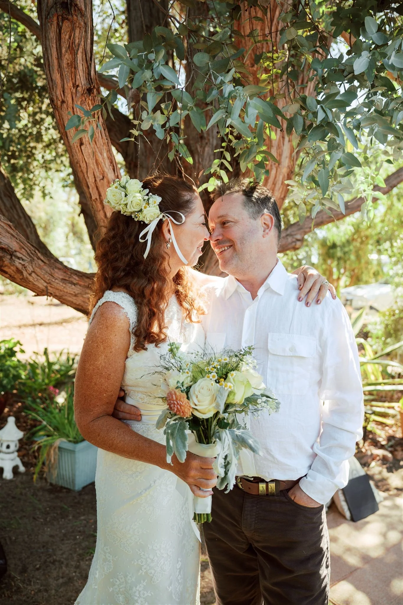A bride and groom smiling and touching foreheads outdoors under a large tree, with the bride holding a bouquet of white roses and greenery.