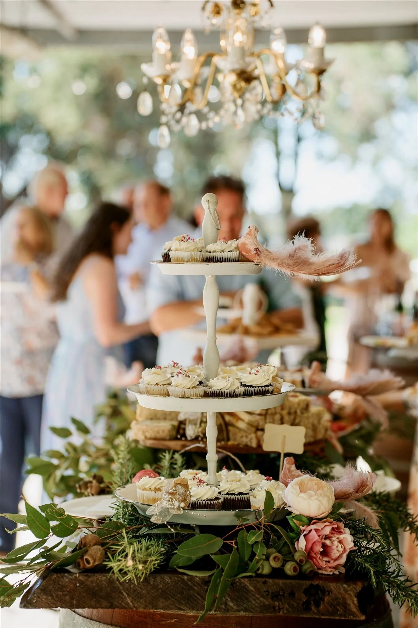 A three-tiered dessert stand with cupcakes and sweets, decorated with flowers, feathers, and greenery, at a festive gathering with blurred guests in the background and a chandelier overhead.
