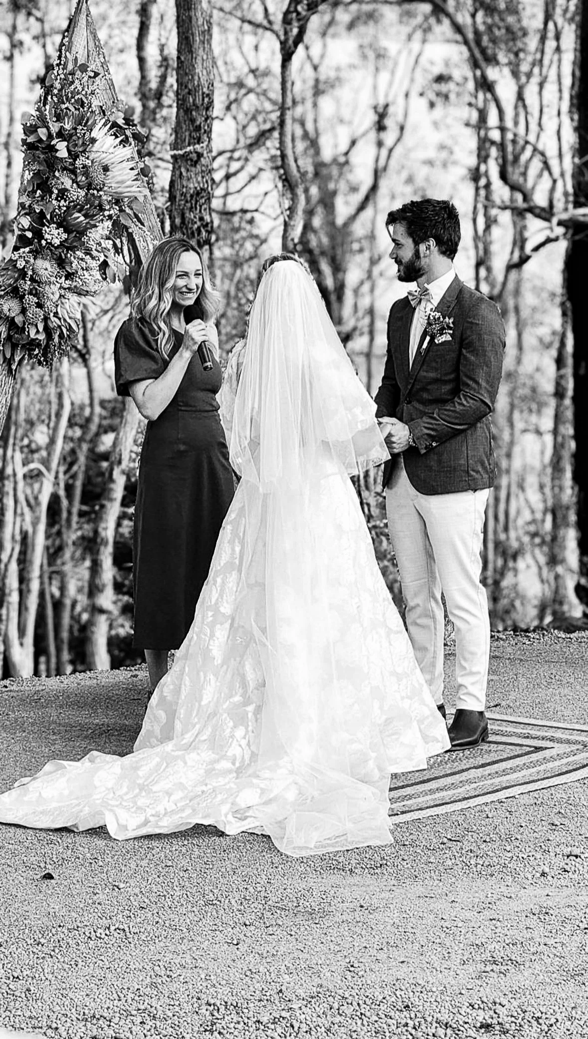 A black and white photo of a wedding ceremony in a forest. A woman in a dark dress is holding a microphone, facing the bride and groom. The bride is wearing a long gown and veil, and the groom is dressed in a suit with a boutonniere. They are holding