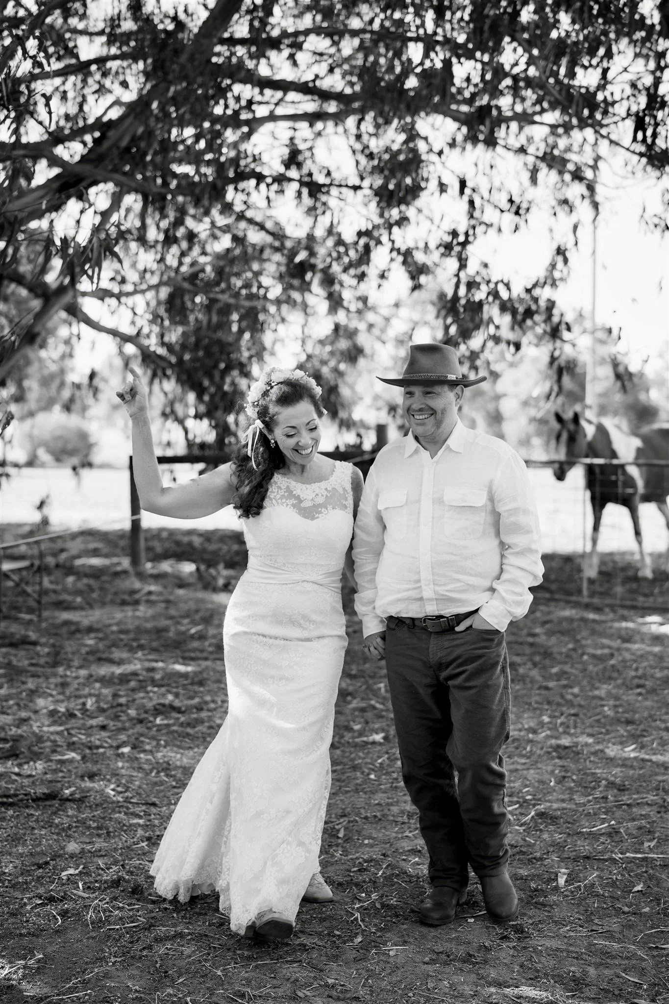 Black and white photo of a bride in a wedding dress and a groom in a cowboy hat standing on farm ground, smiling and holding hands, with trees and horses in the background.