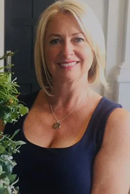 A smiling woman with shoulder-length blonde hair wearing a navy sleeveless top, standing near a potted plant in a room with white walls.