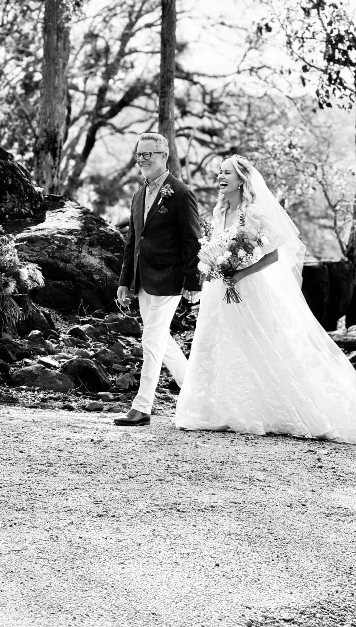 A black and white photo of a smiling bride with her father walking towards her husband to be, outdoors among trees and rocks, with the bride holding a bouquet of flowers.