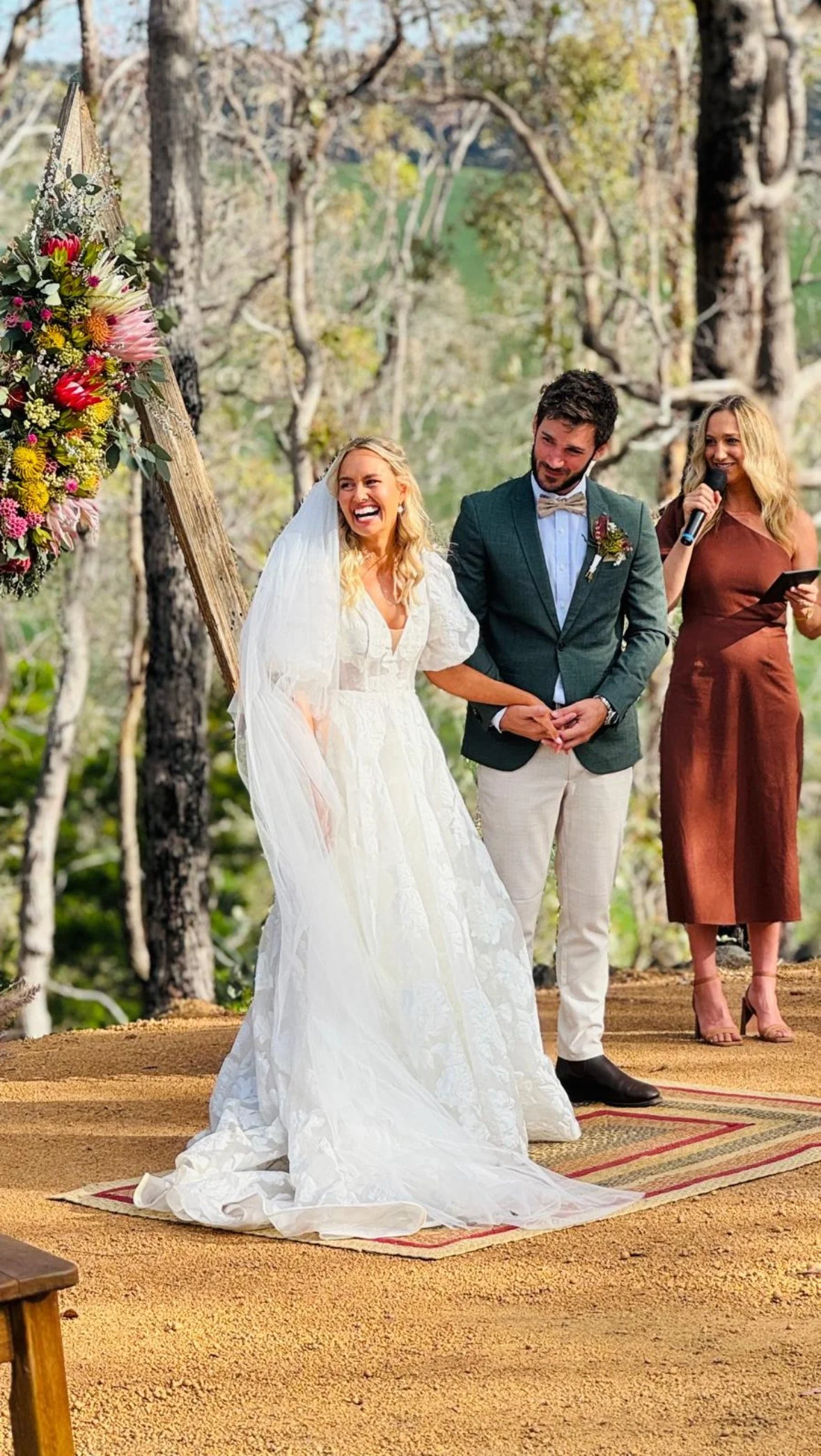 A bride and groom standing hand in hand during their outdoor wedding ceremony, with a officiant woman holding a microphone beside them, and a floral arrangement on a wooden stand behind them in a wooded area.