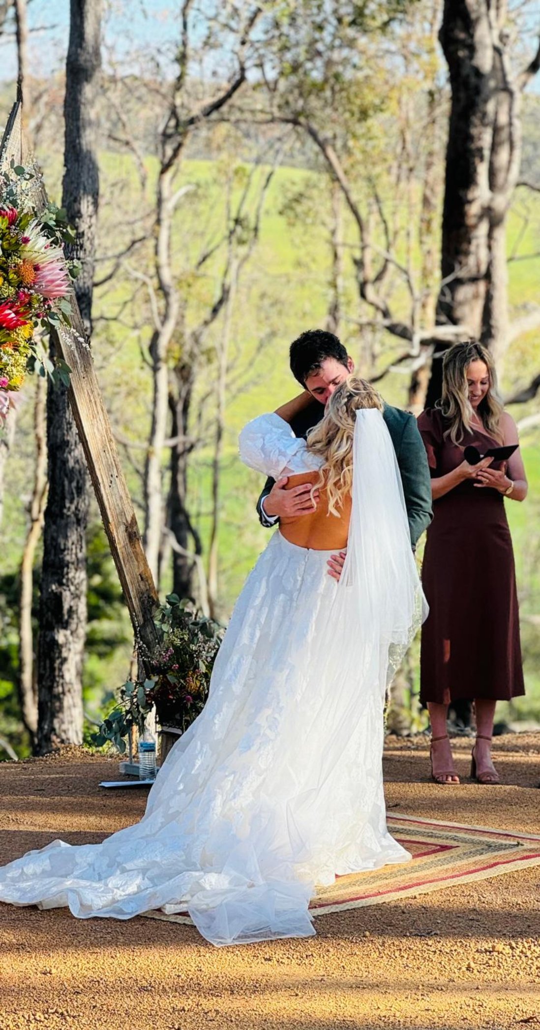 A couple is hugging during an outdoor wedding ceremony in a wooded area, with a woman officiant standing nearby holding a microphone and reading from a card.