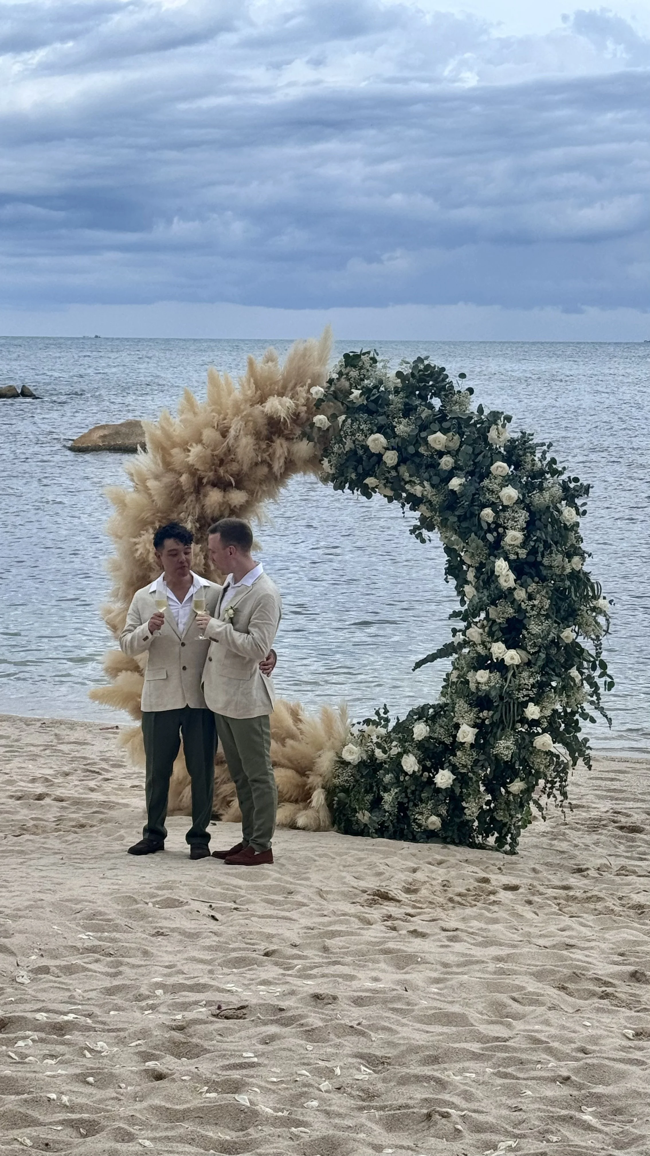 Two men in light-colored suits celebrating with drinks on a beach, standing beside a floral arch decorated with white roses and greenery, with the ocean and cloudy sky in the background.