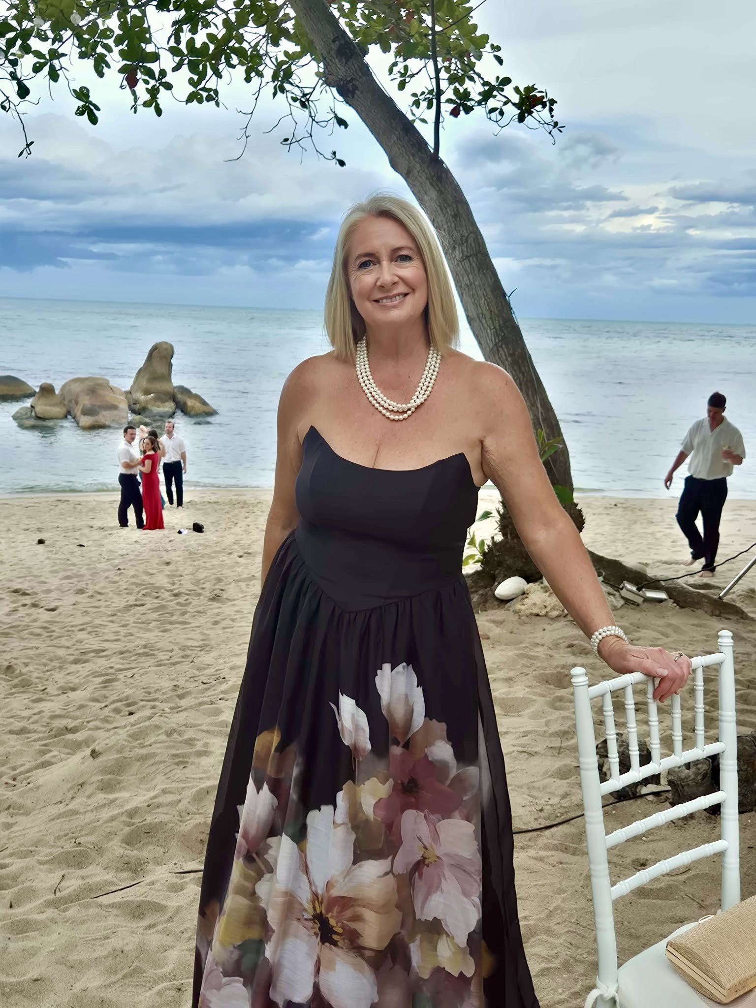 A woman in a strapless black dress with a floral skirt is smiling and standing on a beach. She is wearing multi-strand pearl necklaces and a pearl bracelet. In the background, there are groups of people dressed formally, some near the shoreline, with