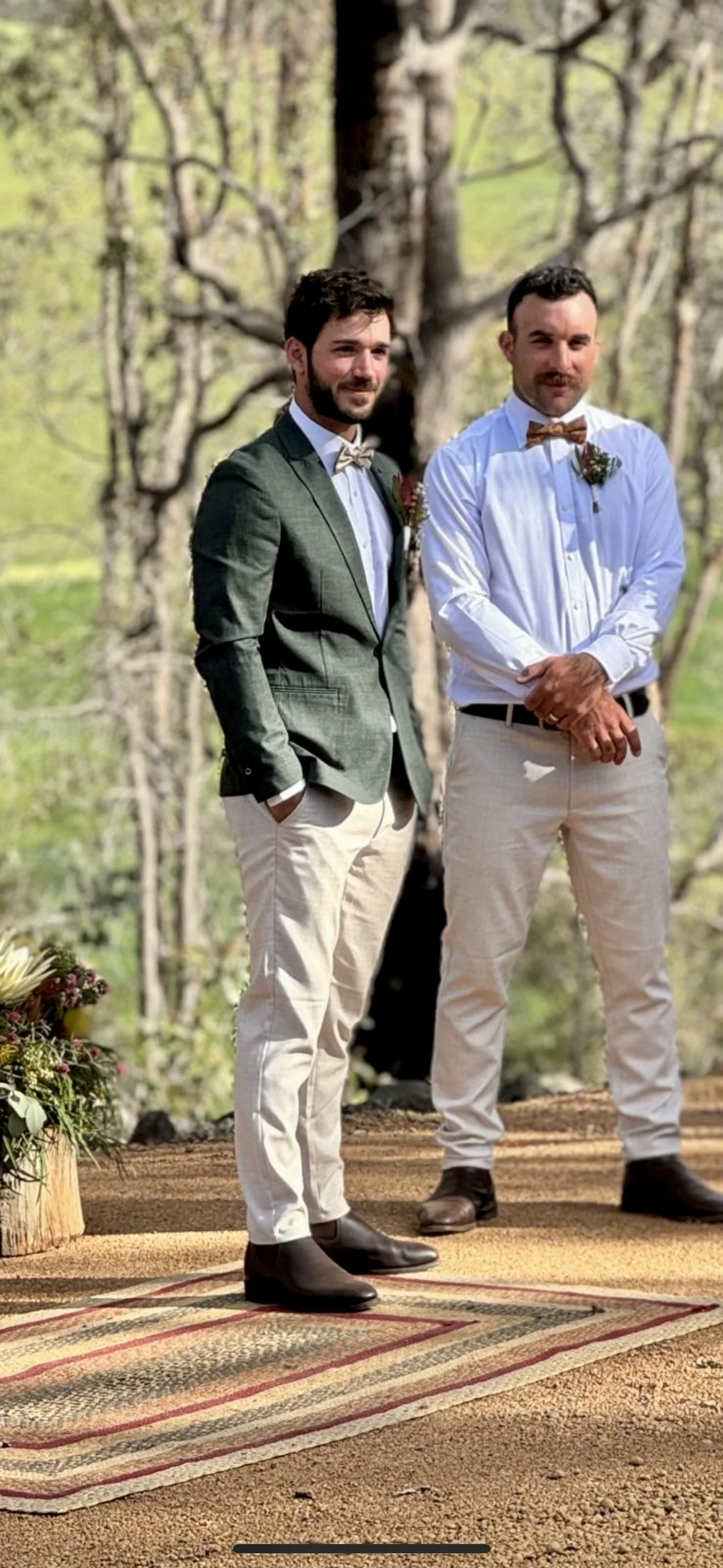 Groom and best man in formal attire standing outdoors on a wedding or ceremony stage, with a large tree in the background and a floral arrangement on the ground.