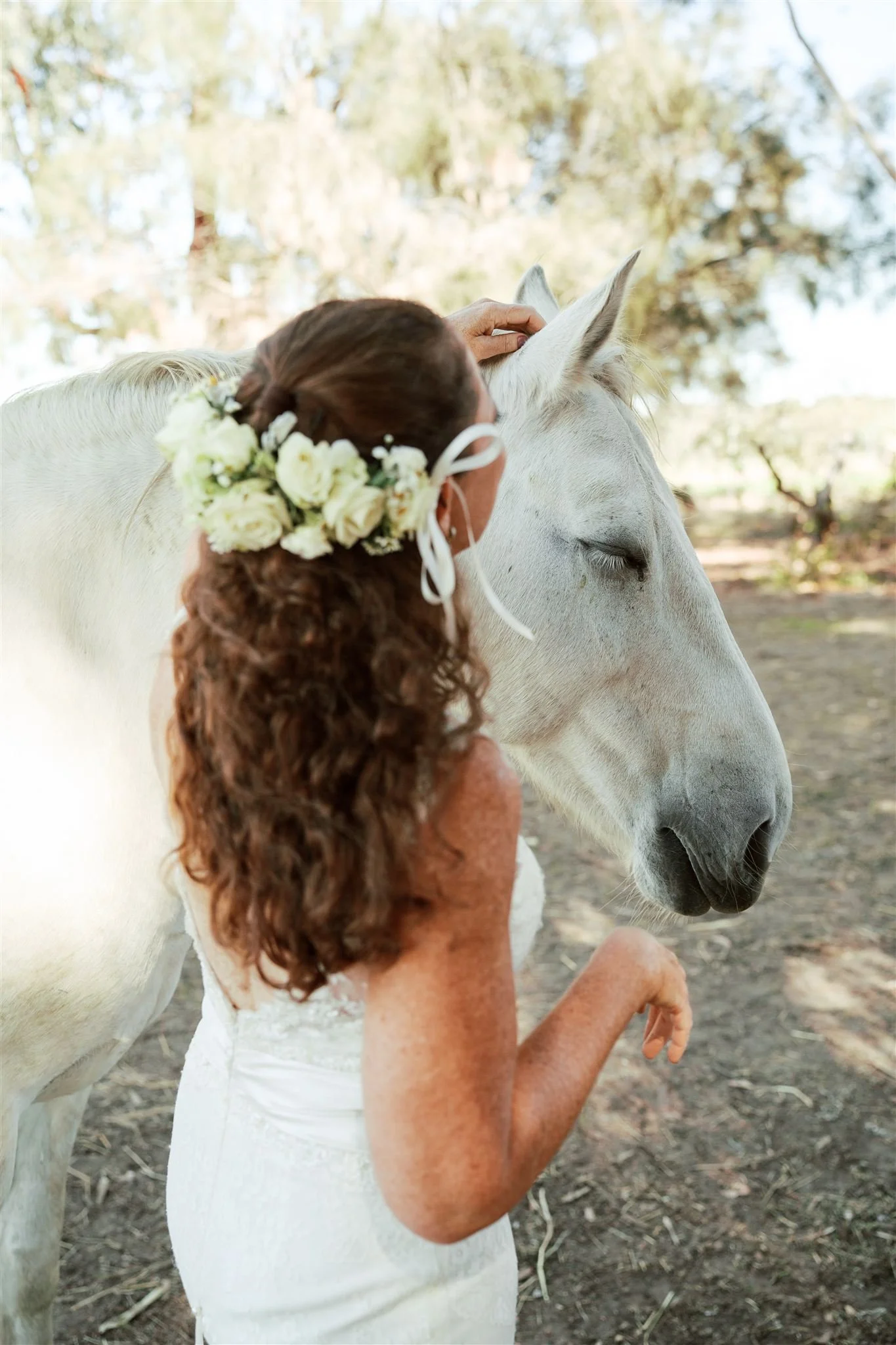 A bride with curly hair wearing a flower crown and white dress gently petting a white horse outdoors in a natural setting.