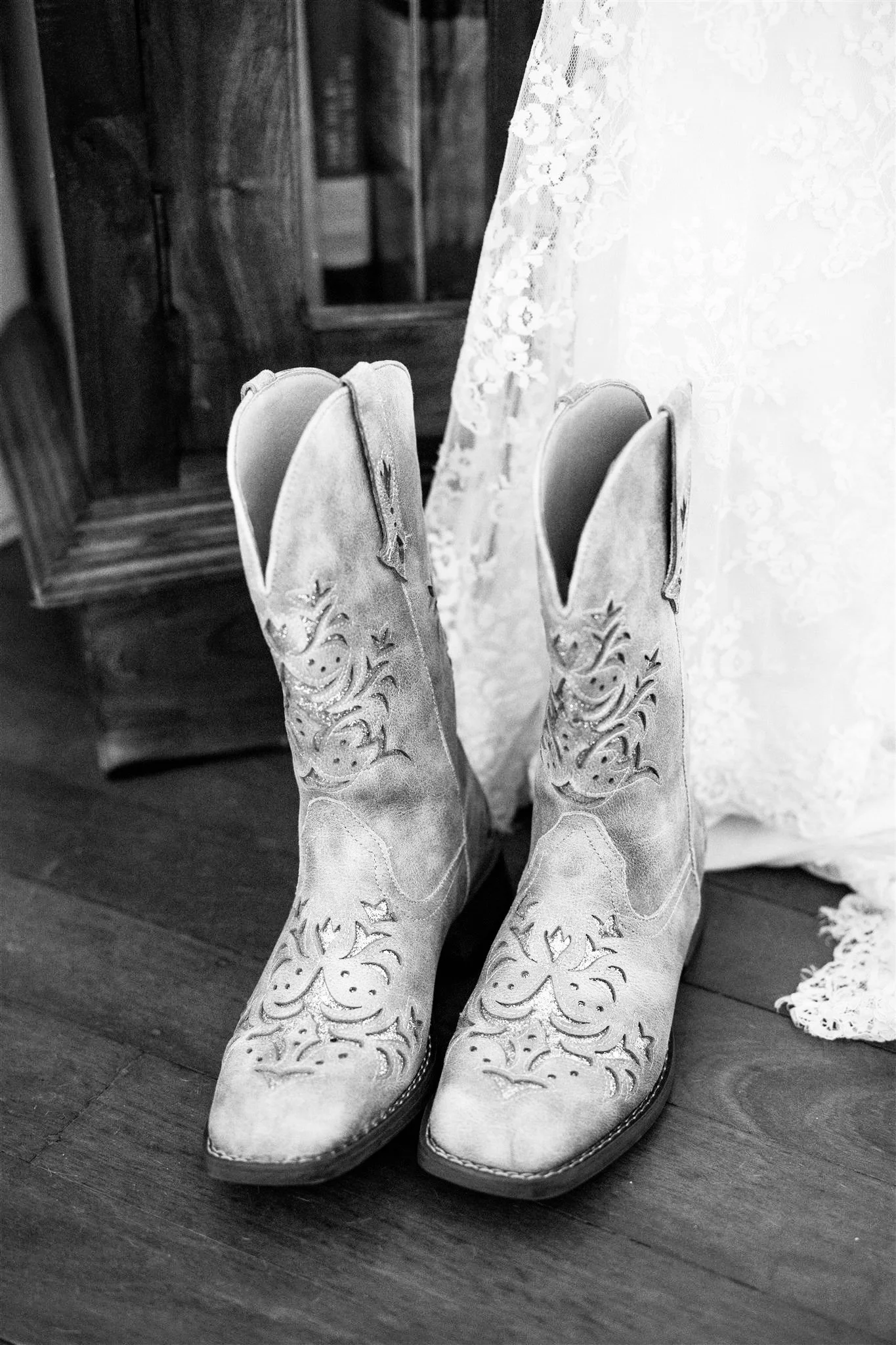 A pair of decorative cowboy boots on a wooden floor next to a lace curtain, with a wooden cabinet in the background.