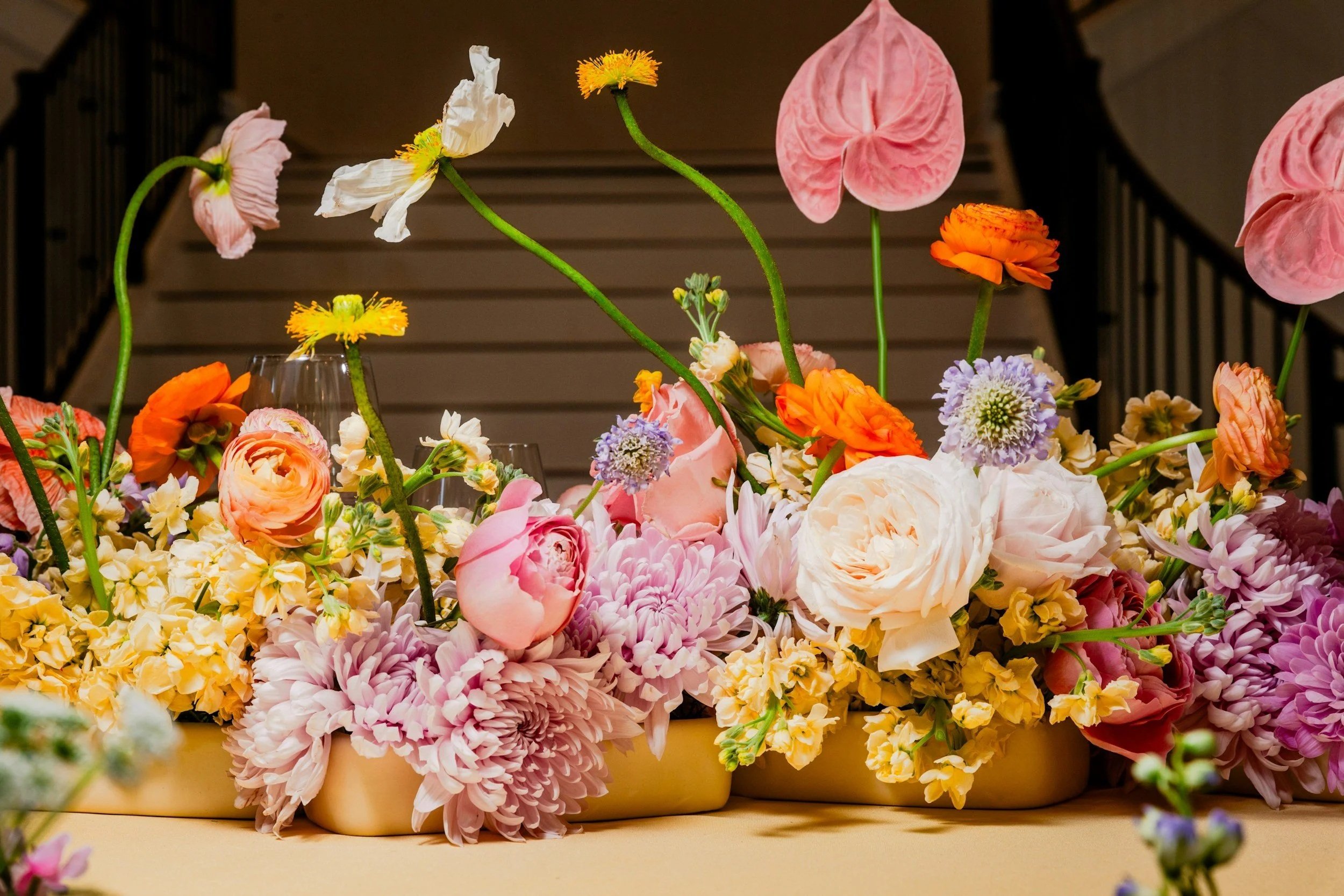 Arrangement of colorful flowers, including pink roses, peonies, orange ranunculus, and other mixed blooms, placed on a yellow surface with stairs in the background.