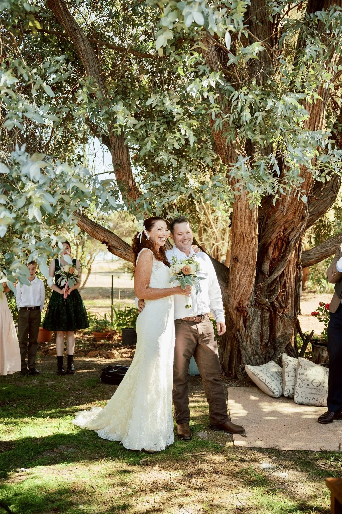 A bride and groom smiling under a large tree at an outdoor wedding ceremony, with guests in the background.