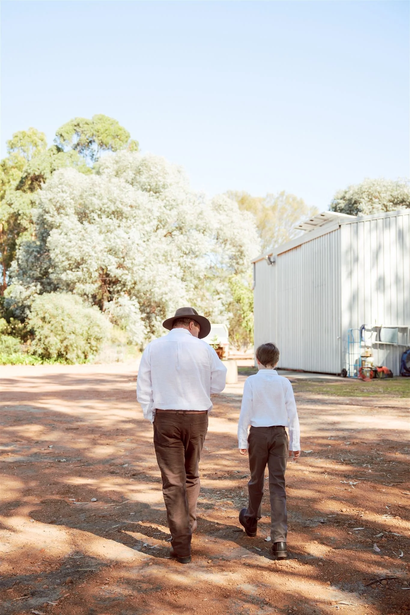 A groom and his son walk away from the camera on a dirt path, dressed in white shirts and dark pants, with trees and a white building in the background on a sunny day.
