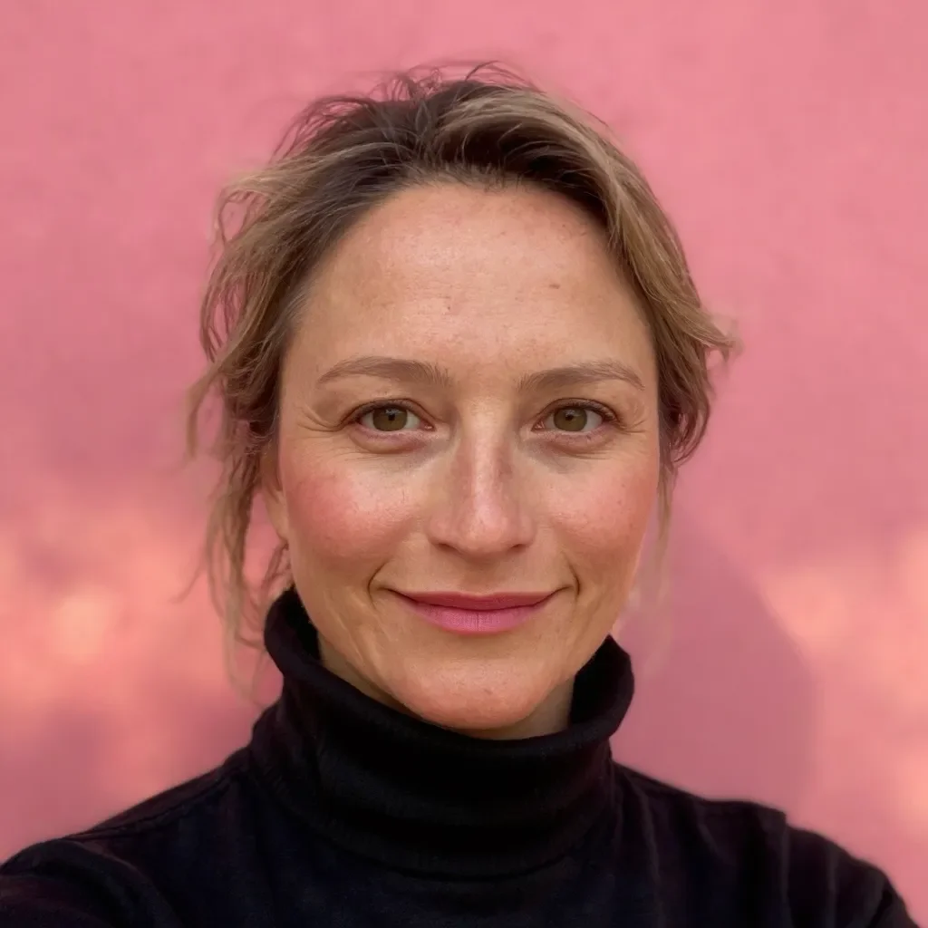 Close-up of a woman with short, wavy blonde hair wearing a black turtleneck, smiling against a pink background.
