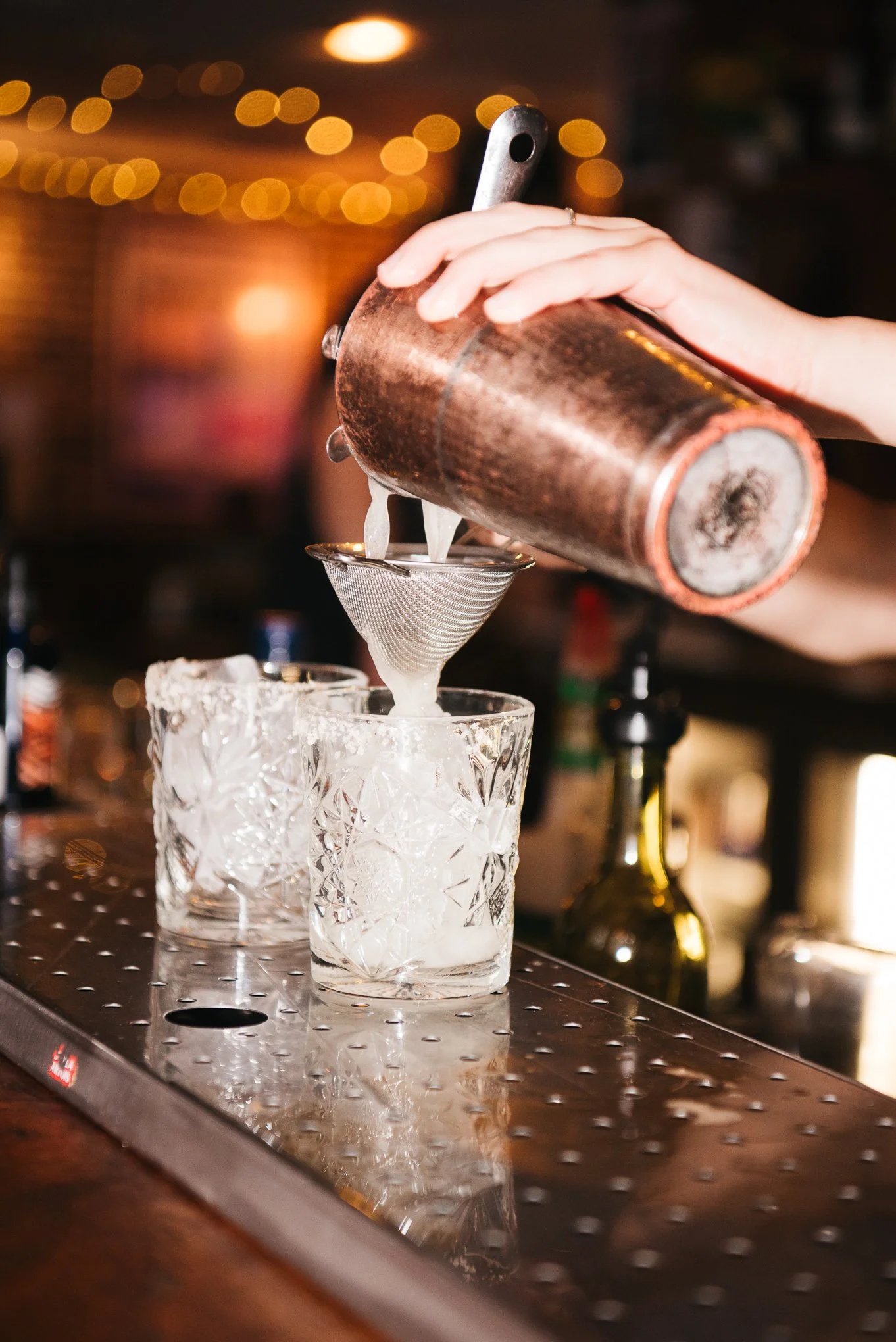 A bartender pouring a cocktail through a strainer into a glass filled with ice at a bar, with warm bokeh lights in the background.