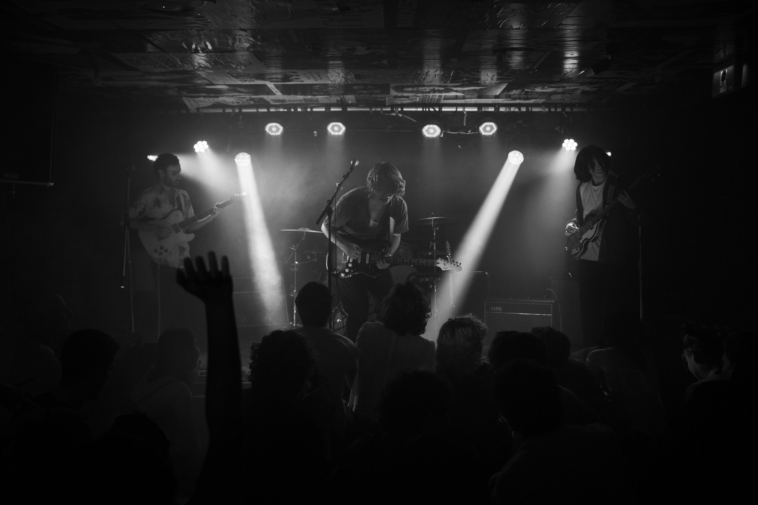 Black and white photo of a live music performance on a small stage, featuring three guitarists playing their instruments with an audience in front of them. The stage is lit with spotlights.