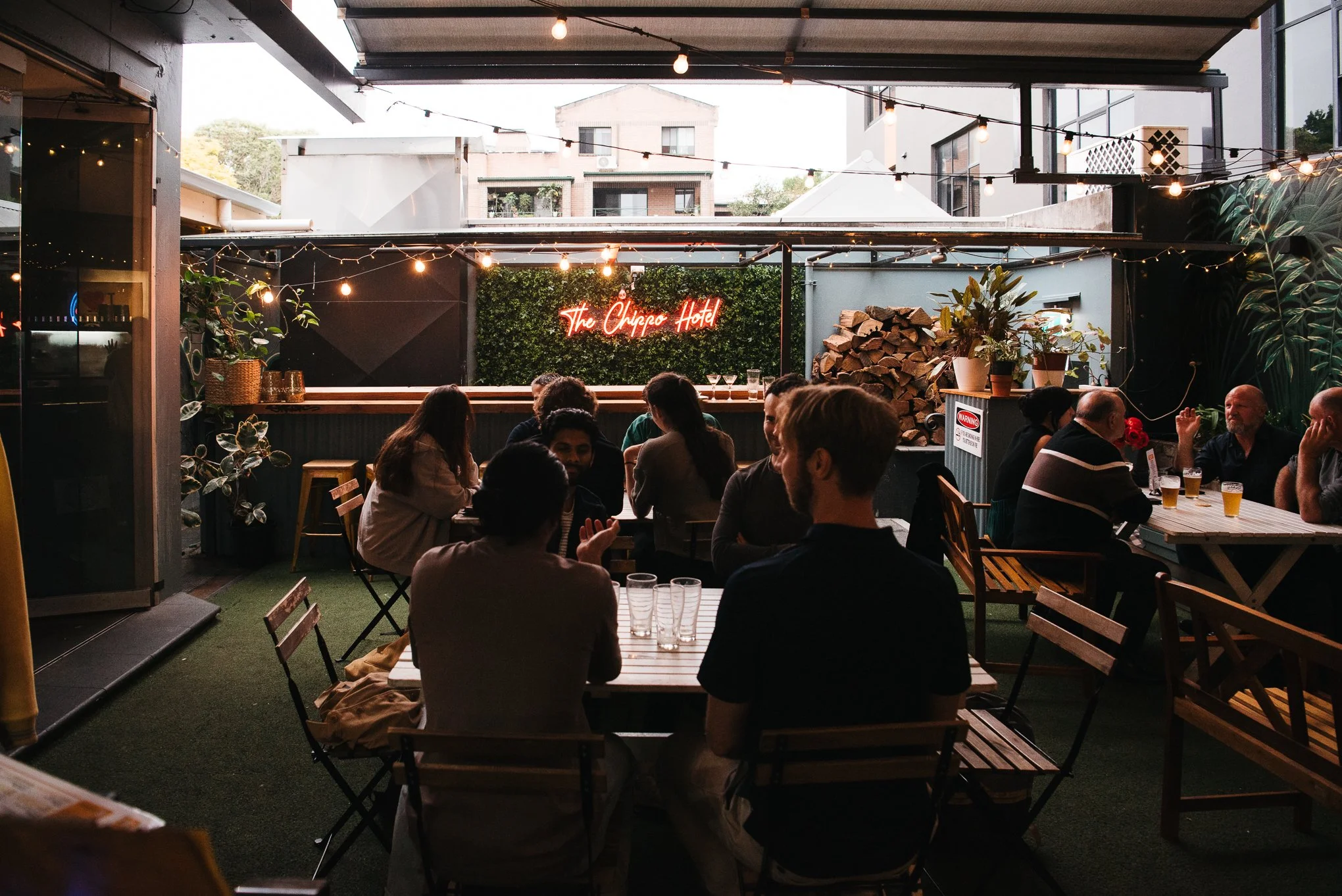 People dining at an outdoor patio with string lights, a green wall with a neon sign that reads 'The Chippo Hotel,' and stacked firewood.