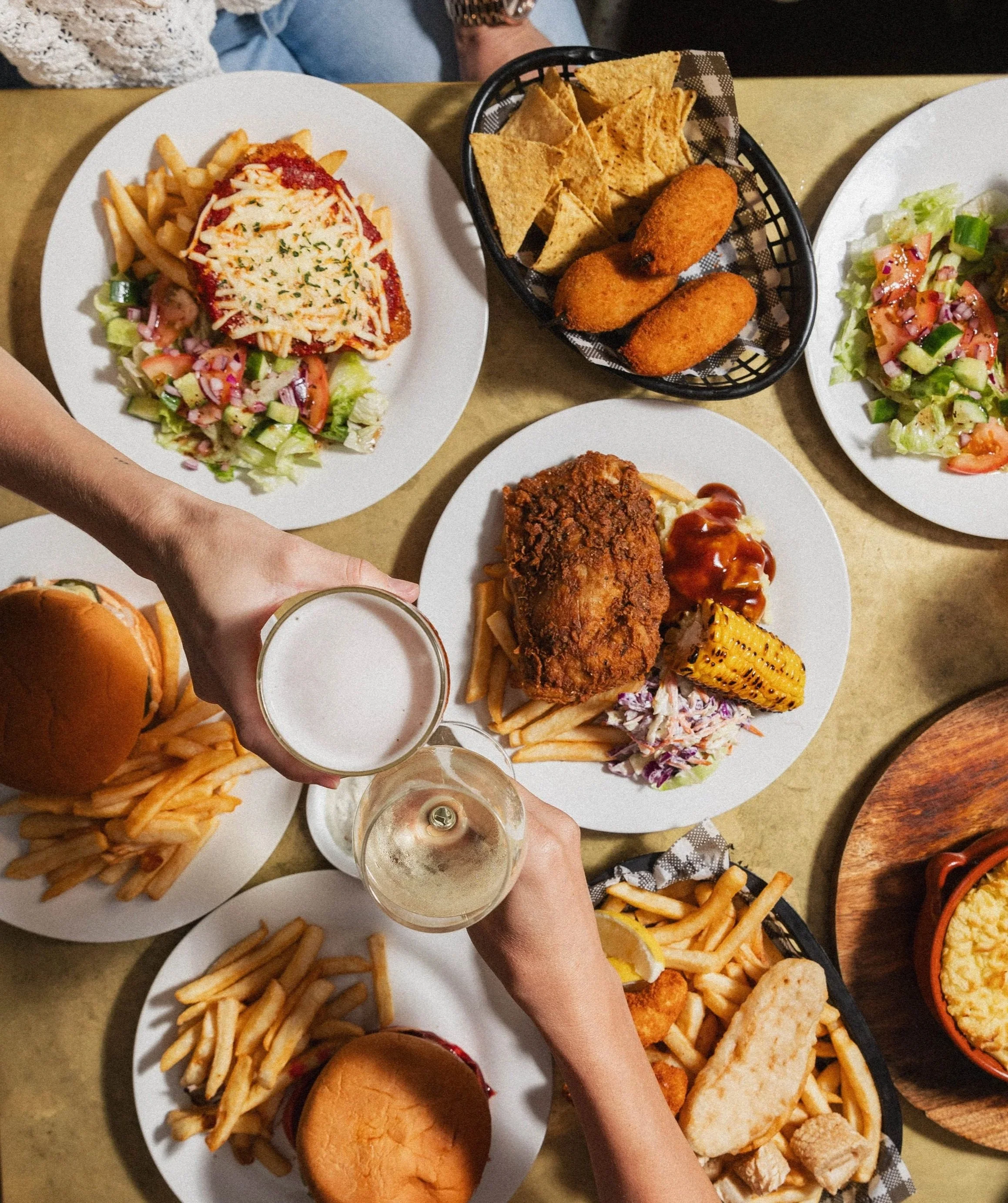 Top-down view of a table with various plates of food, including a burger, fries, salad, fried chicken with ketchup, grilled corn, tortilla chips, mozzarella sticks, and drinks being clinked in a toast.
