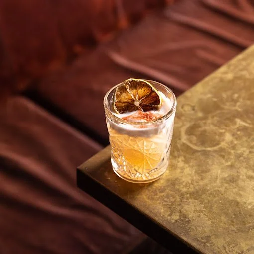 A glass with a clear beverage and a dried lime wedge garnish on top, placed on a table with a marble surface.