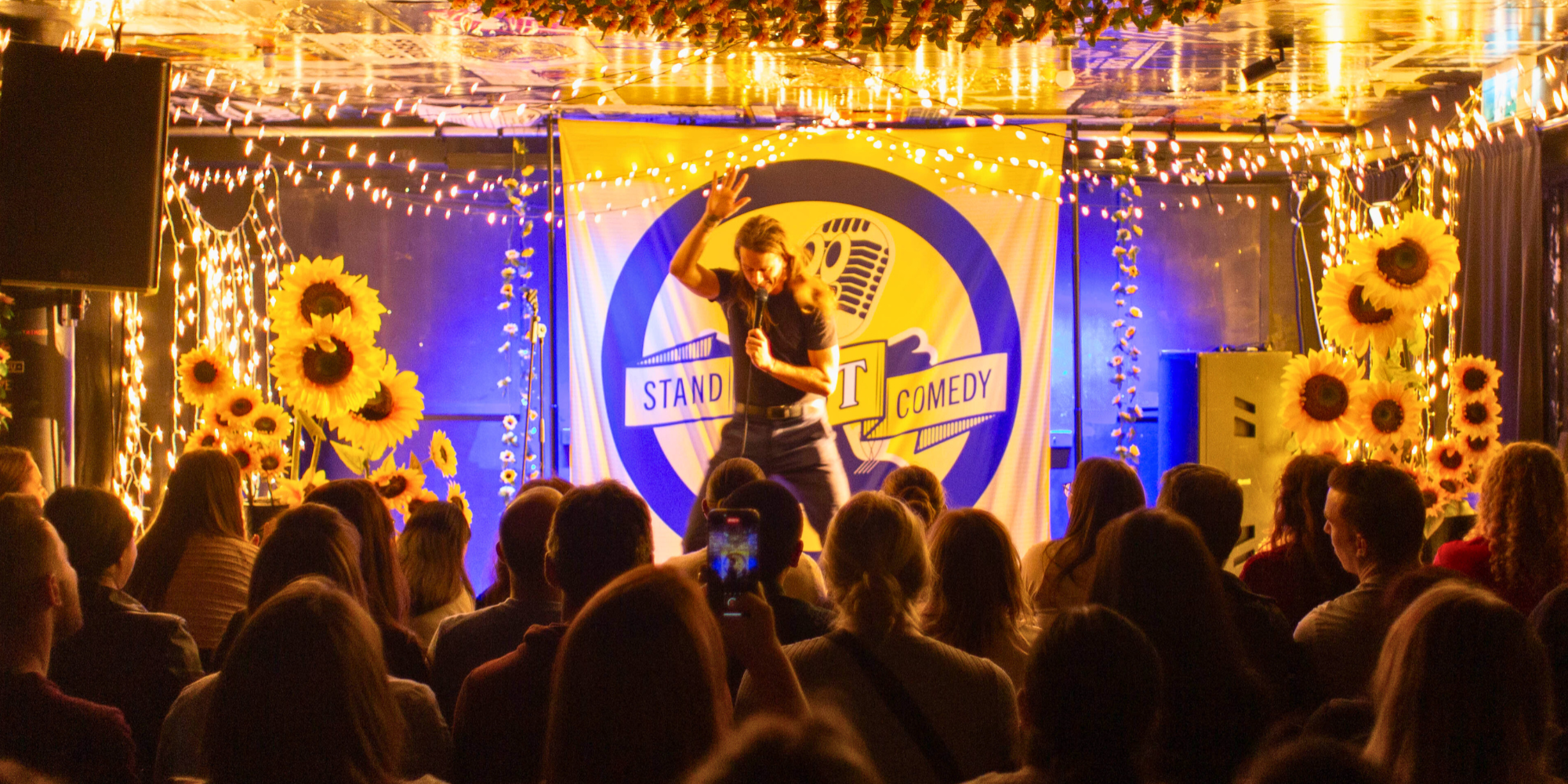 Female stand-up comedian performing on stage with an audience in a decorated comedy club, featuring sunflower arrangements, string lights, and a banner with 'Stand Up Comedy' on it.