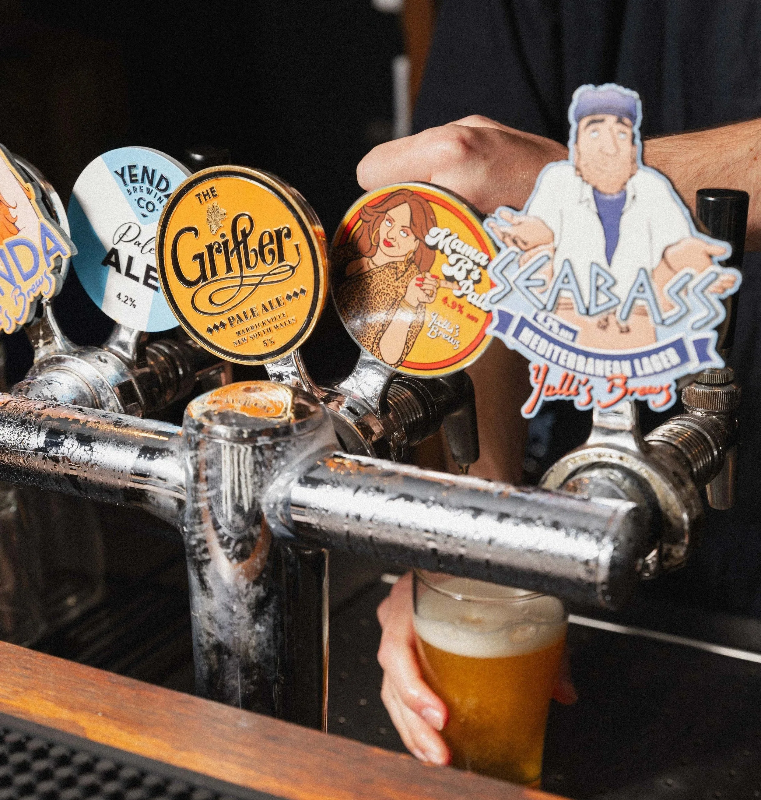 A person pouring beer from tap handles at a bar. The tap handles have colorful labels, including one for Grifter Pale Ale, Yield Beer, and Skullbass Lager. The person is holding a glass of beer underneath the taps.