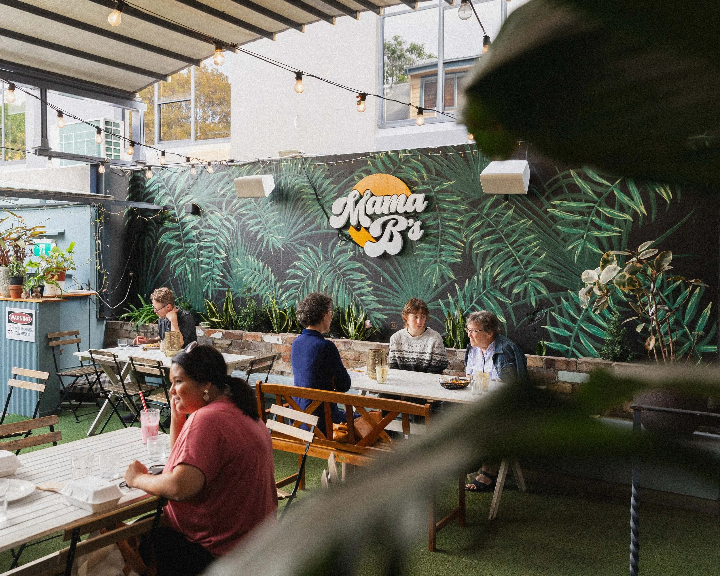 People dining at an outdoor patio of a restaurant with a tropical plant mural and string lights, with a sign reading 'Mama B' in the background.