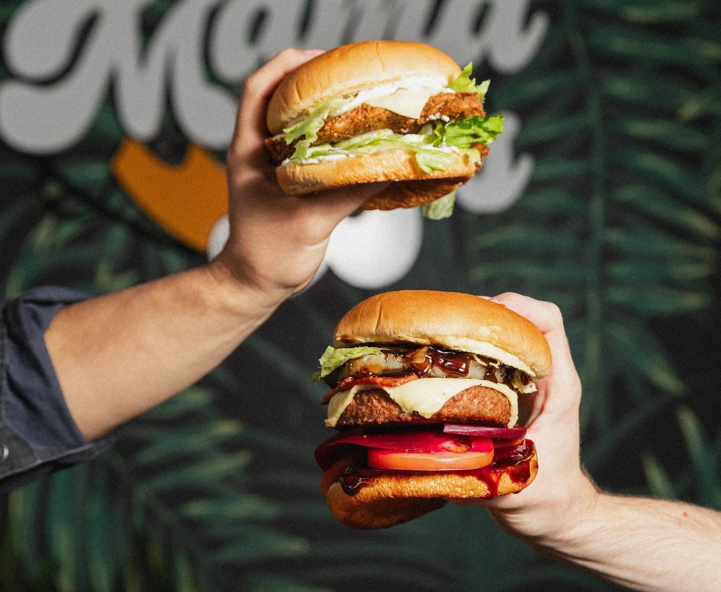 Two hands holding large burgers with lettuce, tomato, cheese, and various toppings against a leafy background.