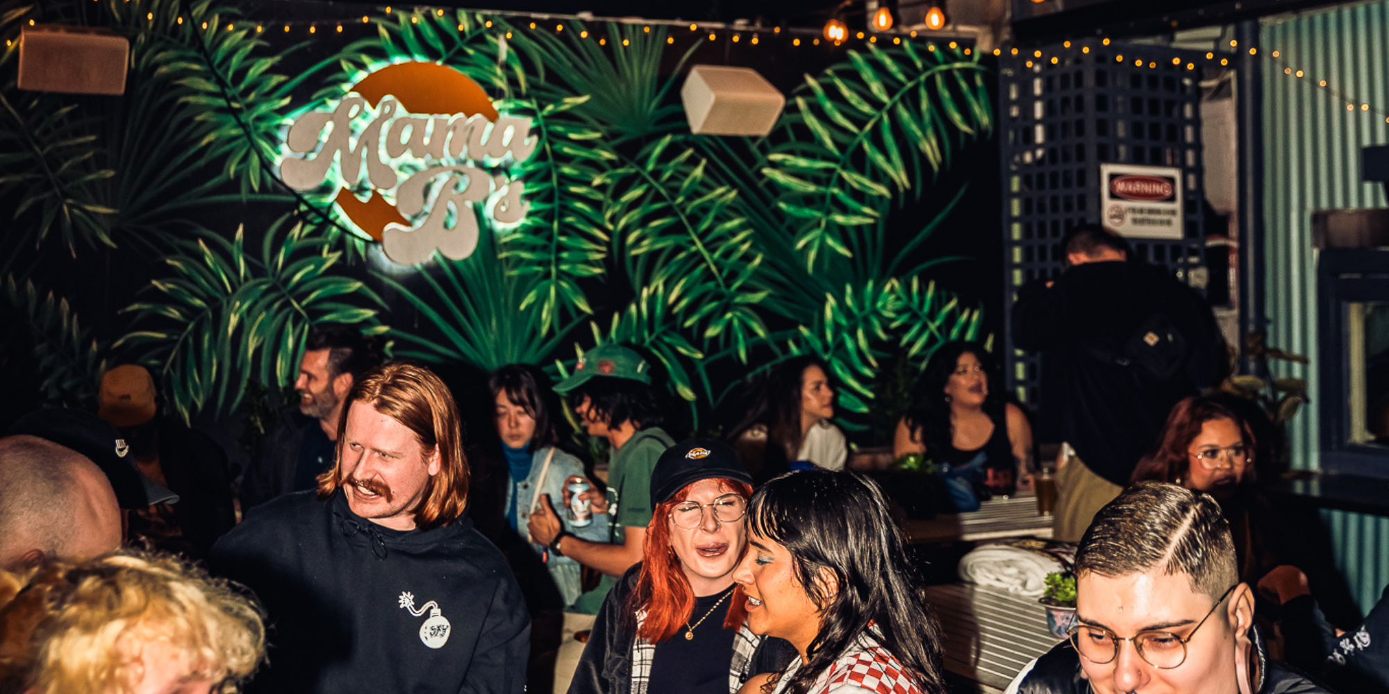 People socializing at a crowded indoor party with tropical decor and a neon sign that says 'Mama B'.
