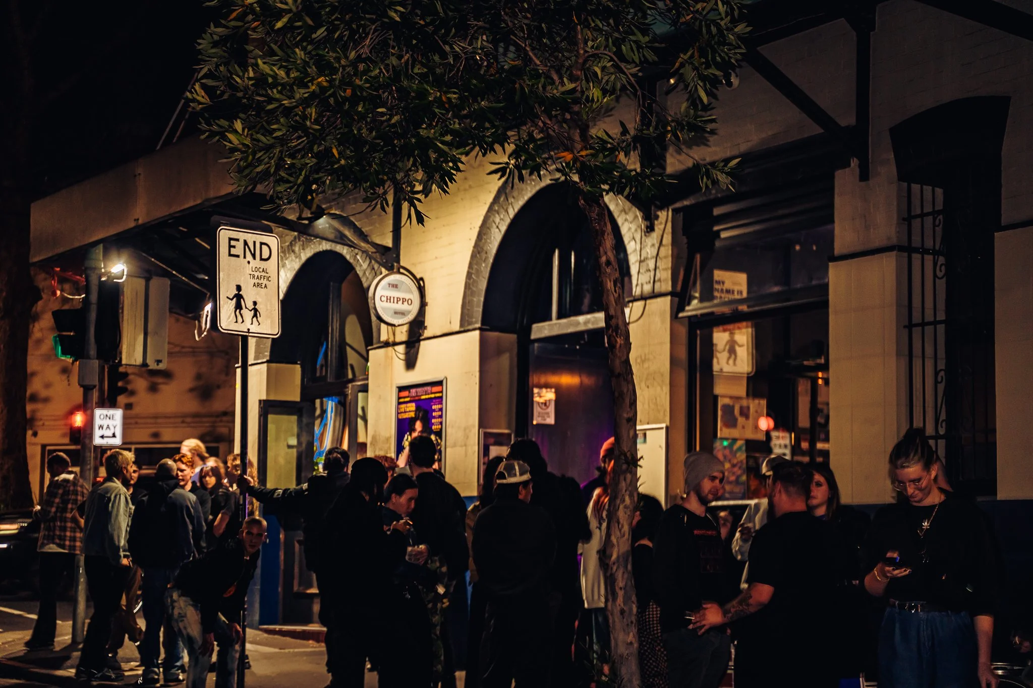 People waiting outside a venue at night with signs and lights, with trees and a building in the background.