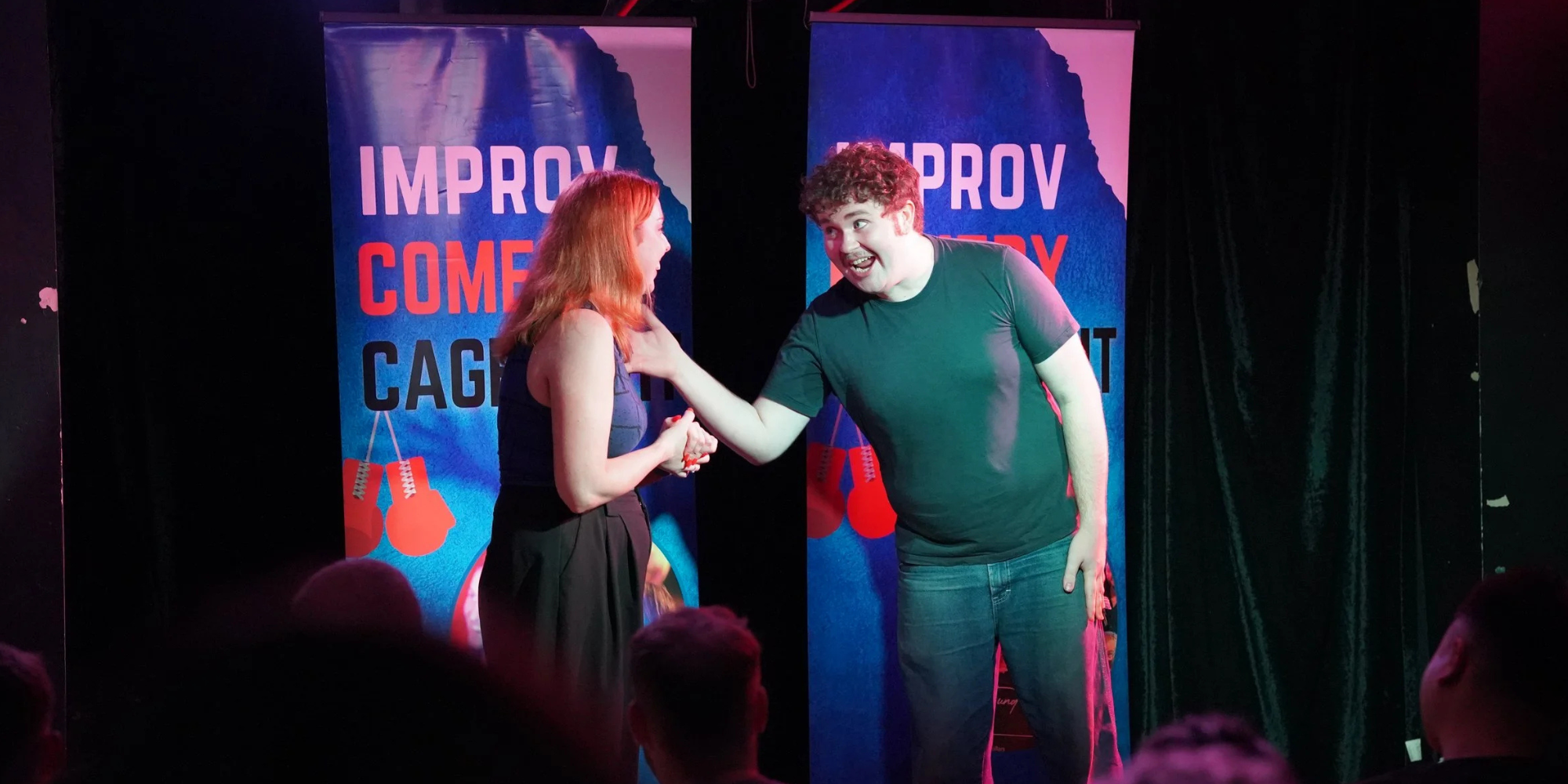 A comedy improv show featuring two performers on stage with black curtains, behind two colorful posters that say 'Improv Comedy Cag'. One performer is a red-haired woman in a dark top and skirt, and the other is a curly-haired man in a black T-shirt and jeans, smiling and engaging with each other.
