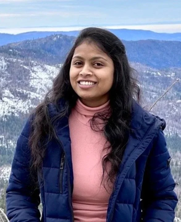 A woman with long dark hair smiling outdoors against a mountainous landscape with snow-capped peaks.